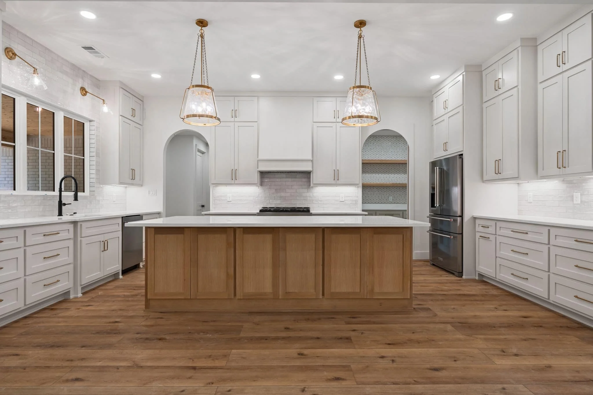 Modern white kitchen with wooden island, white cabinetry, stainless steel refrigerator, black faucet, and wood flooring, illuminated by ceiling and pendant lights.