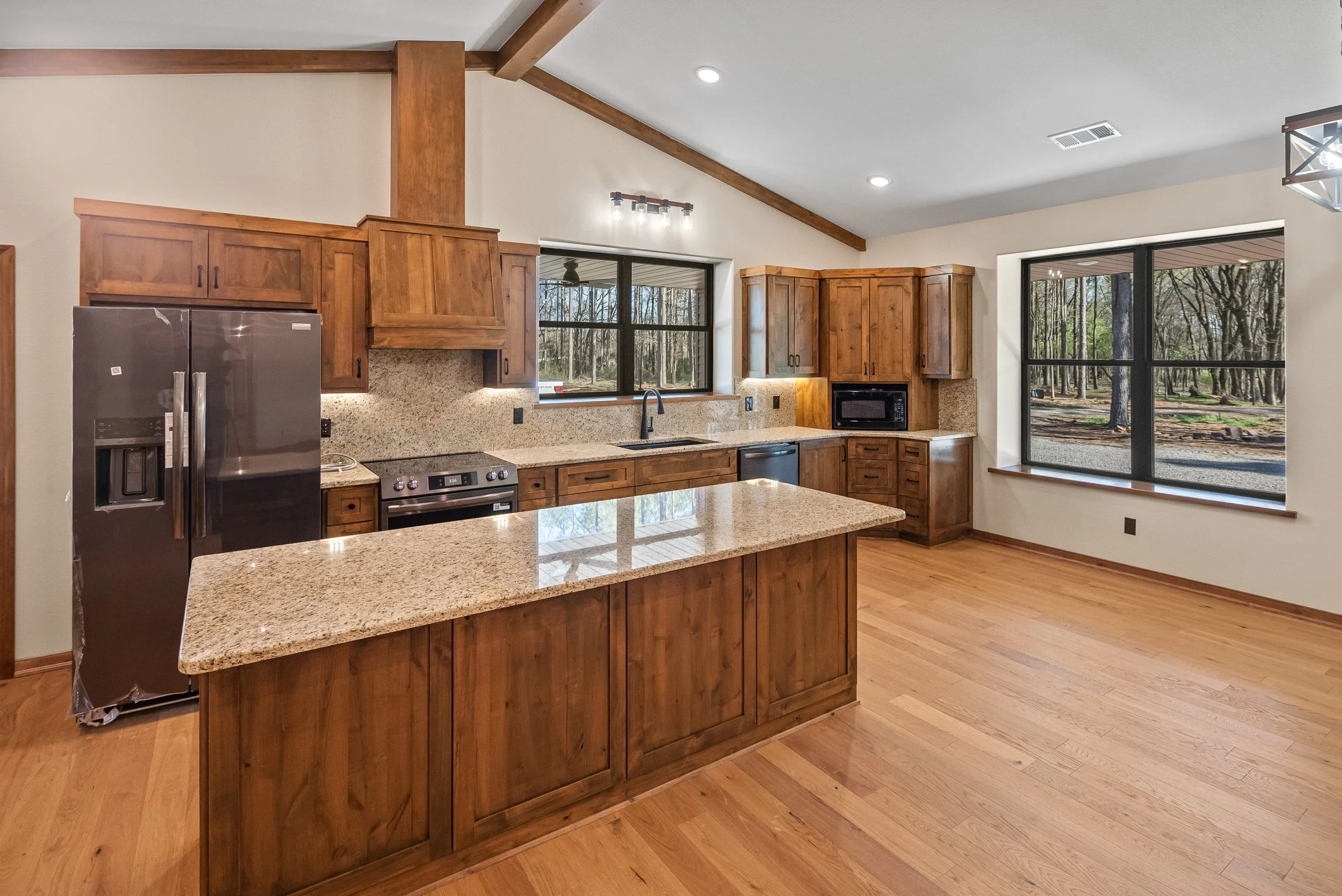 Kitchen with wooden cabinets, granite countertops, stainless steel refrigerator, stove, microwave, and sink, with large windows overlooking a wooded yard.