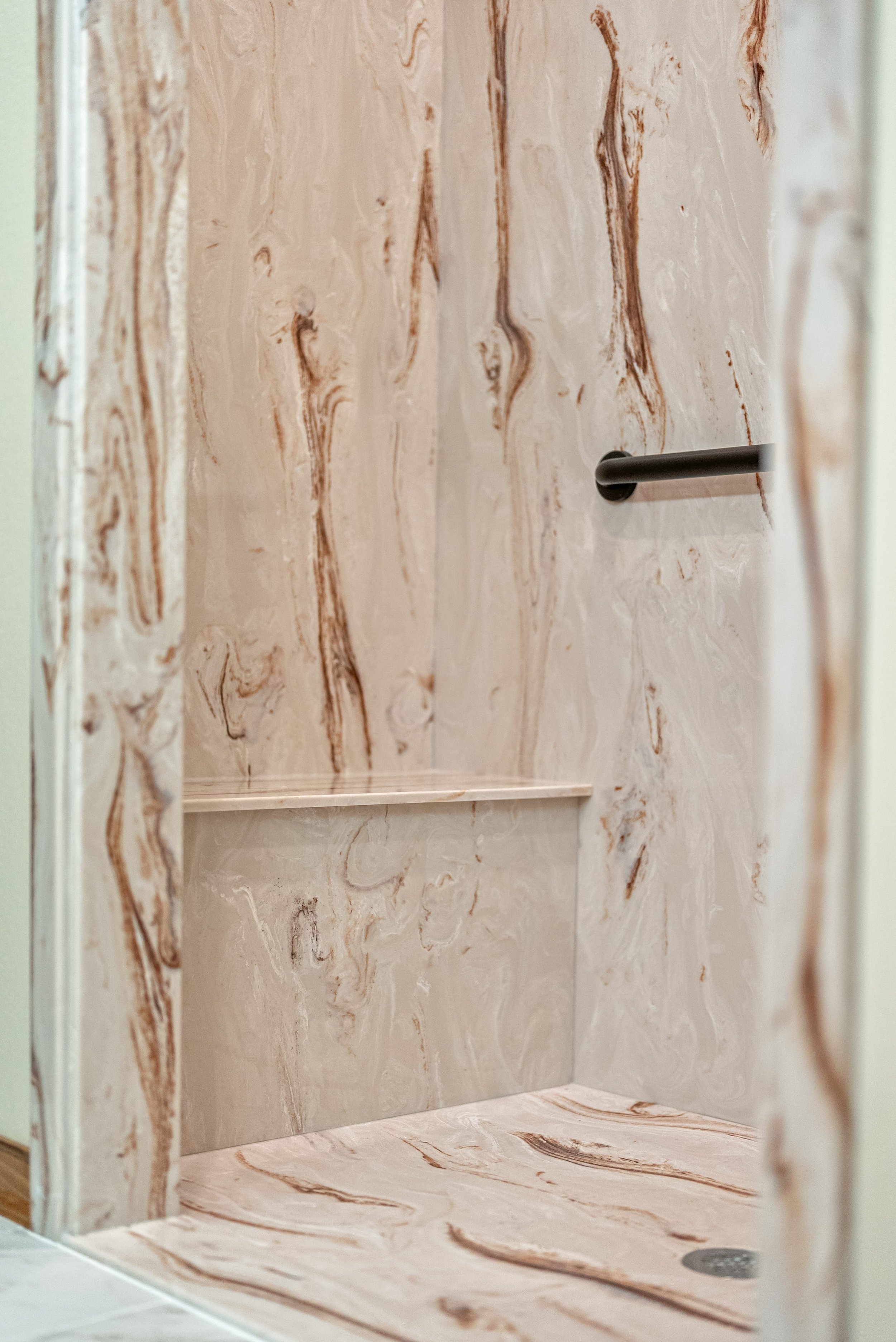 A marble shower with a beige and brown veined pattern, featuring a black grab bar on the right wall and a built-in shelf at the back, with a circular drain on the floor.