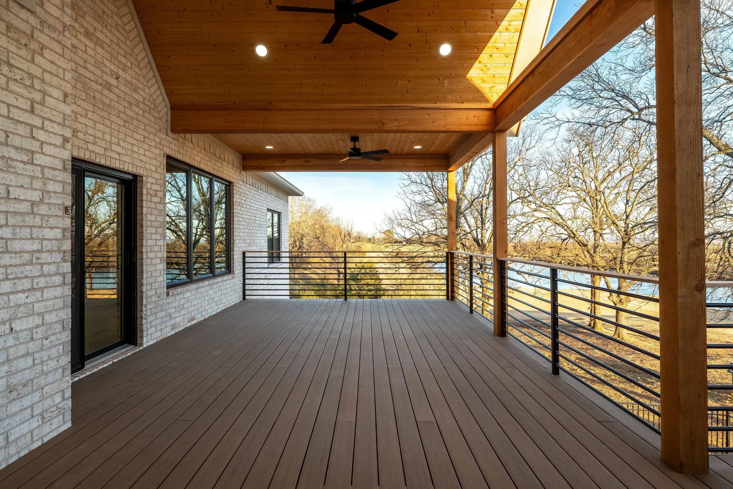 Empty balcony with wooden flooring and ceiling, black metal railing, enclosed by a brick wall with large windows and a door, outdoor trees visible in the background, ceiling fans and recessed lighting installed.