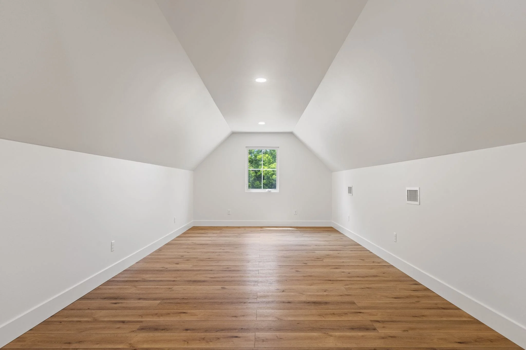 Empty room with white walls, a window showing green trees outside, and natural wood flooring.