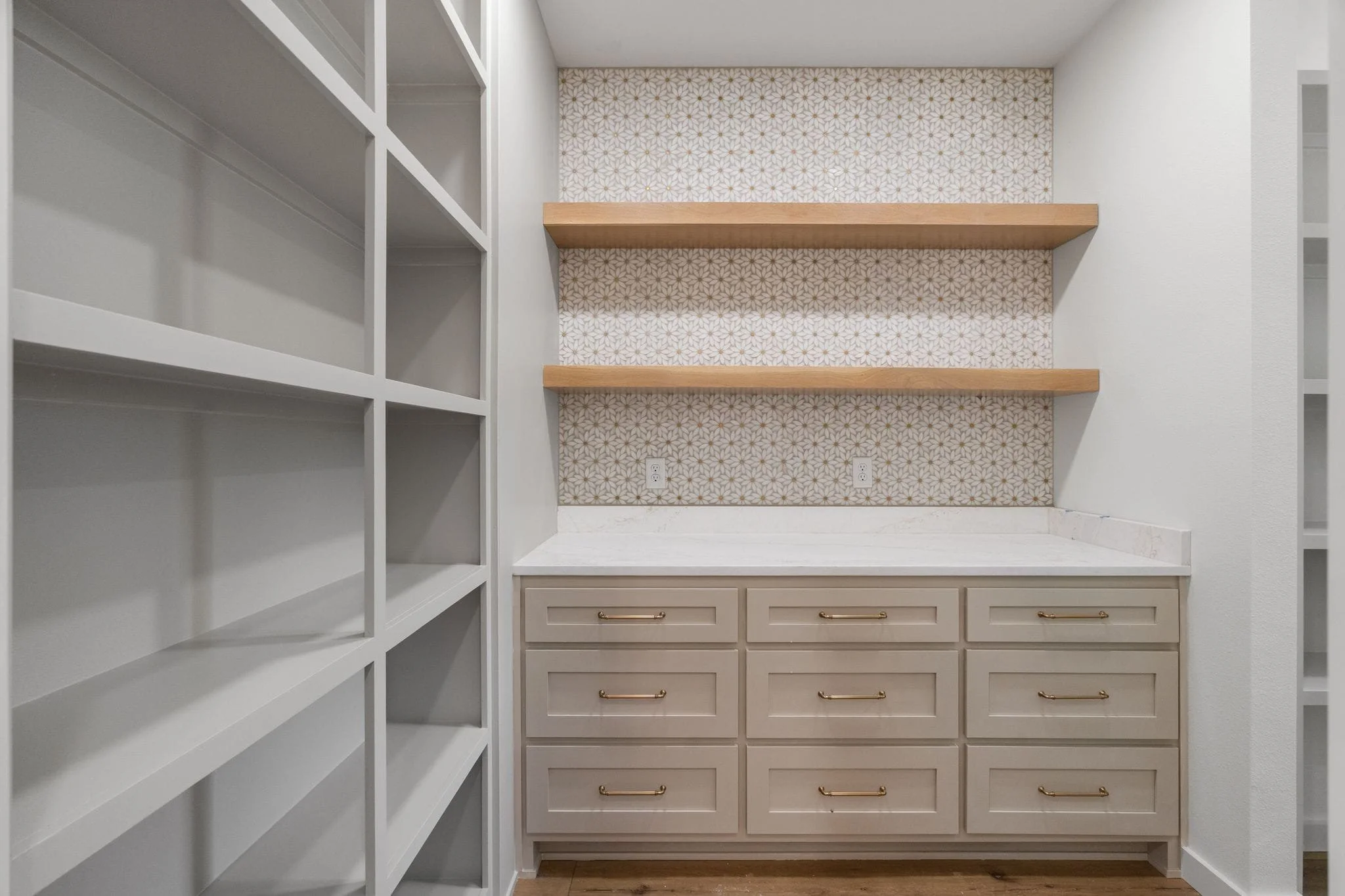 A pantry or storage closet with white cabinetry and drawers at the bottom, two horizontal wooden shelves against a cheery geometric wallpapered back wall, and a white granite countertop below the shelves.