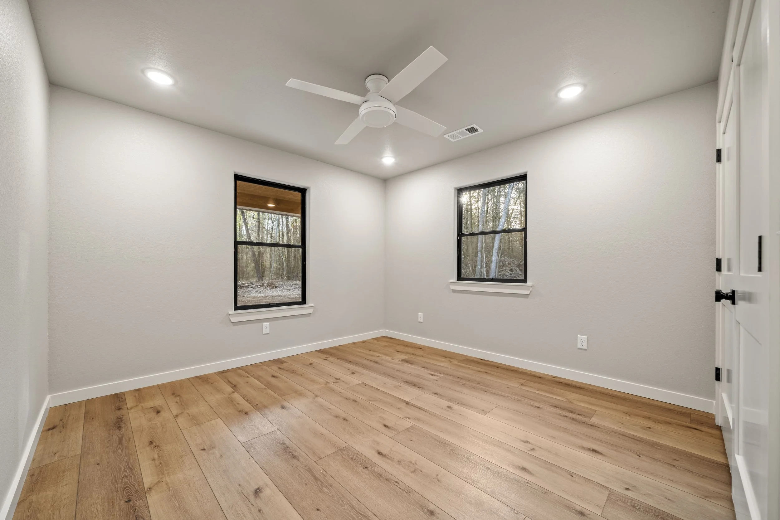 Empty room with white walls, two windows showing a wooded outdoor view, wooden floor, ceiling fan, and ceiling lights.