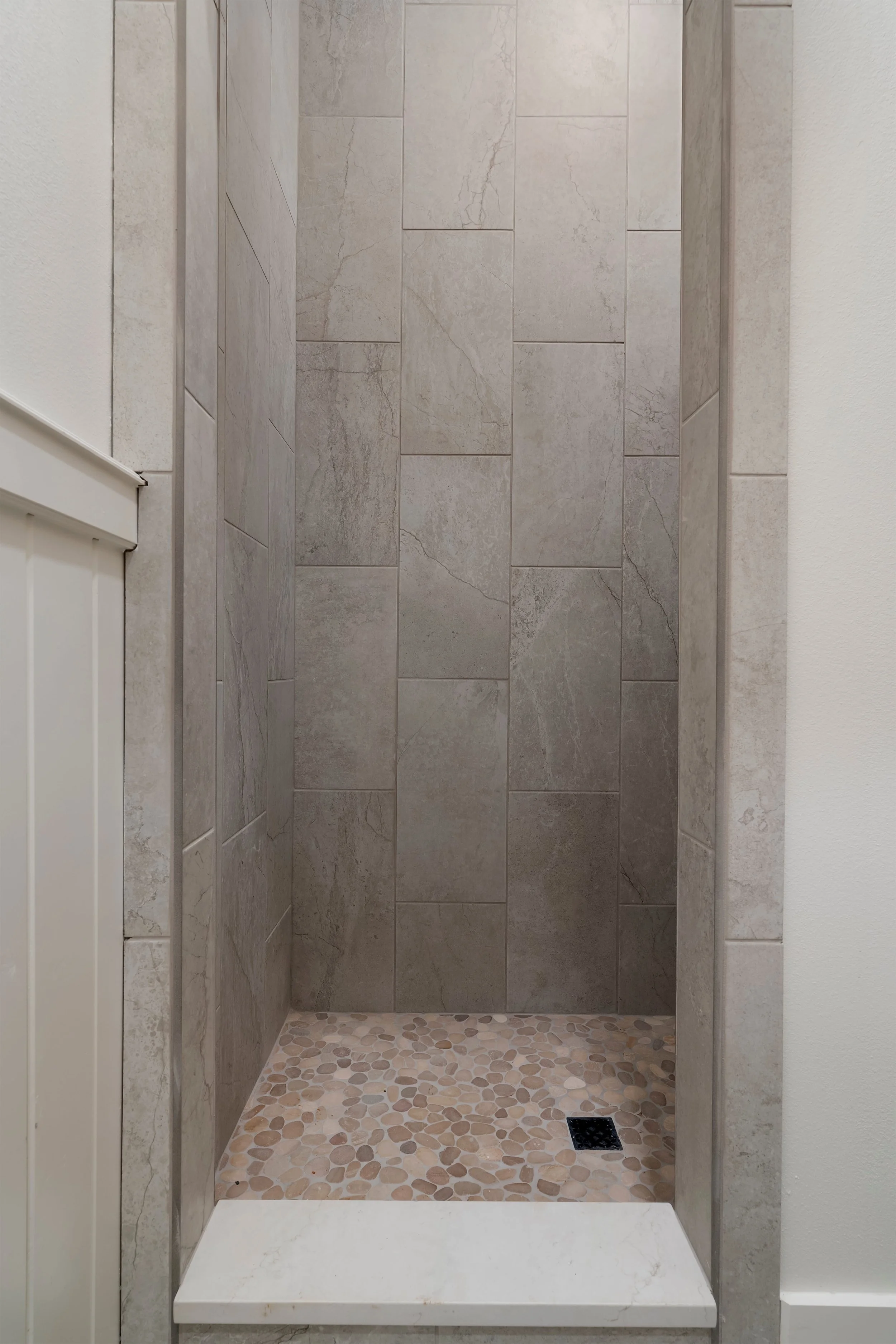Shower area with beige tiles on the walls, a pebble stone floor, and a black drain in the corner.