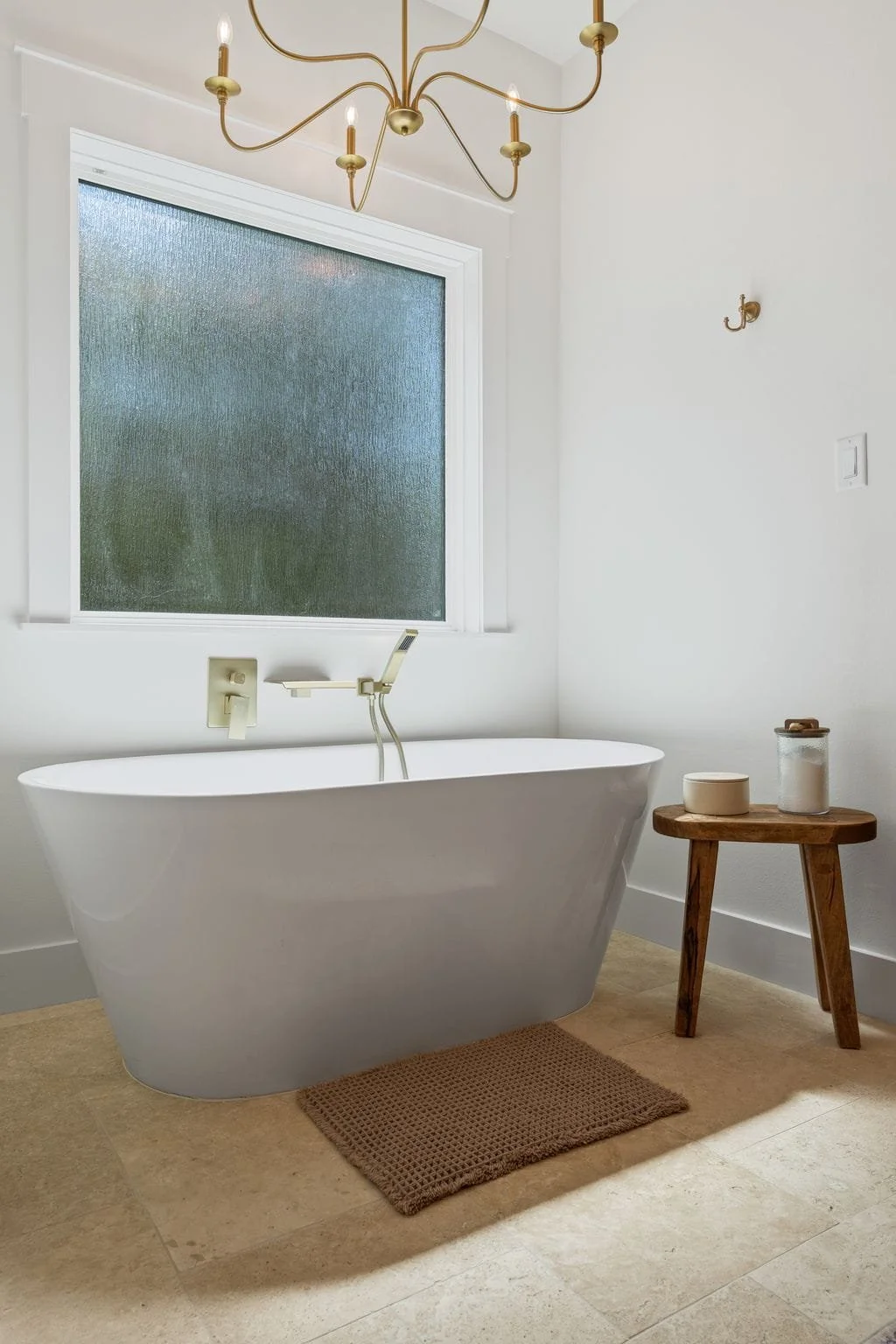 Freestanding soaking tub beneath frosted window with brass fixtures, chandelier lighting, and neutral tile flooring.