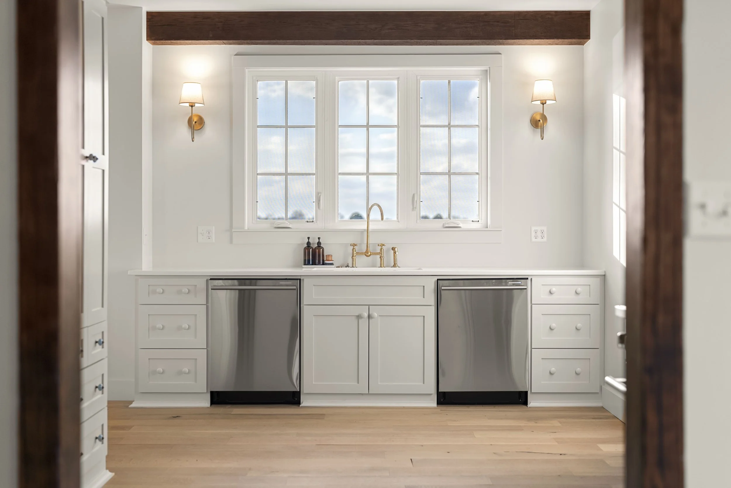 A bright kitchen with a large window, white cabinets, stainless steel dishwasher, brass fixtures, and hardwood floors.