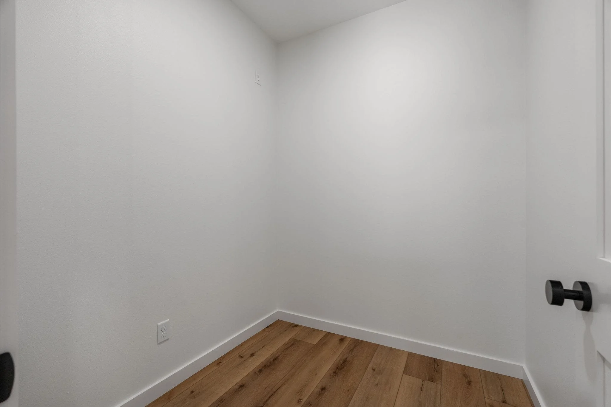 Empty corner of a room with white walls, hardwood floor, electrical outlet, and a black wall hook on the right side.