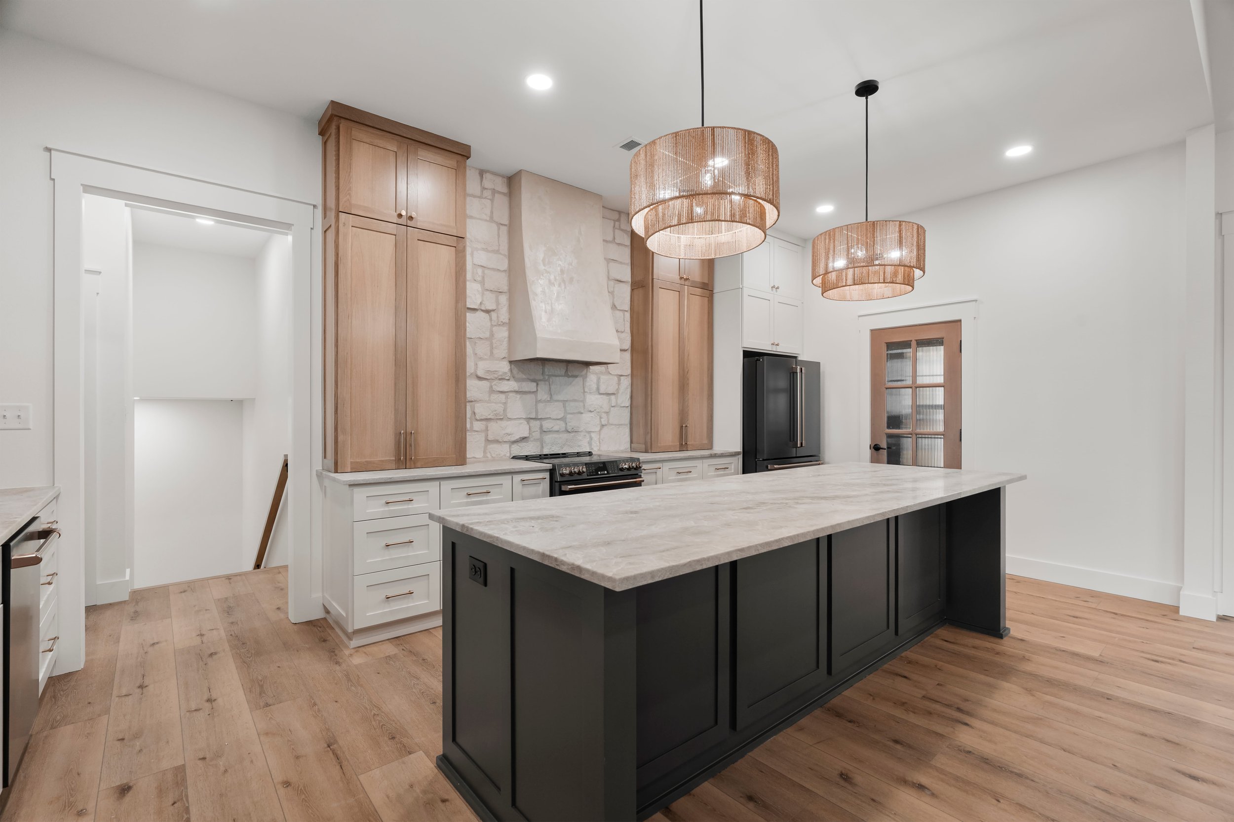 Modern kitchen with light wood and white cabinets, a black island with a cream countertop, stone accents on the wall, a range hood, hardwood flooring, pendant lighting, and a glass-paned door.