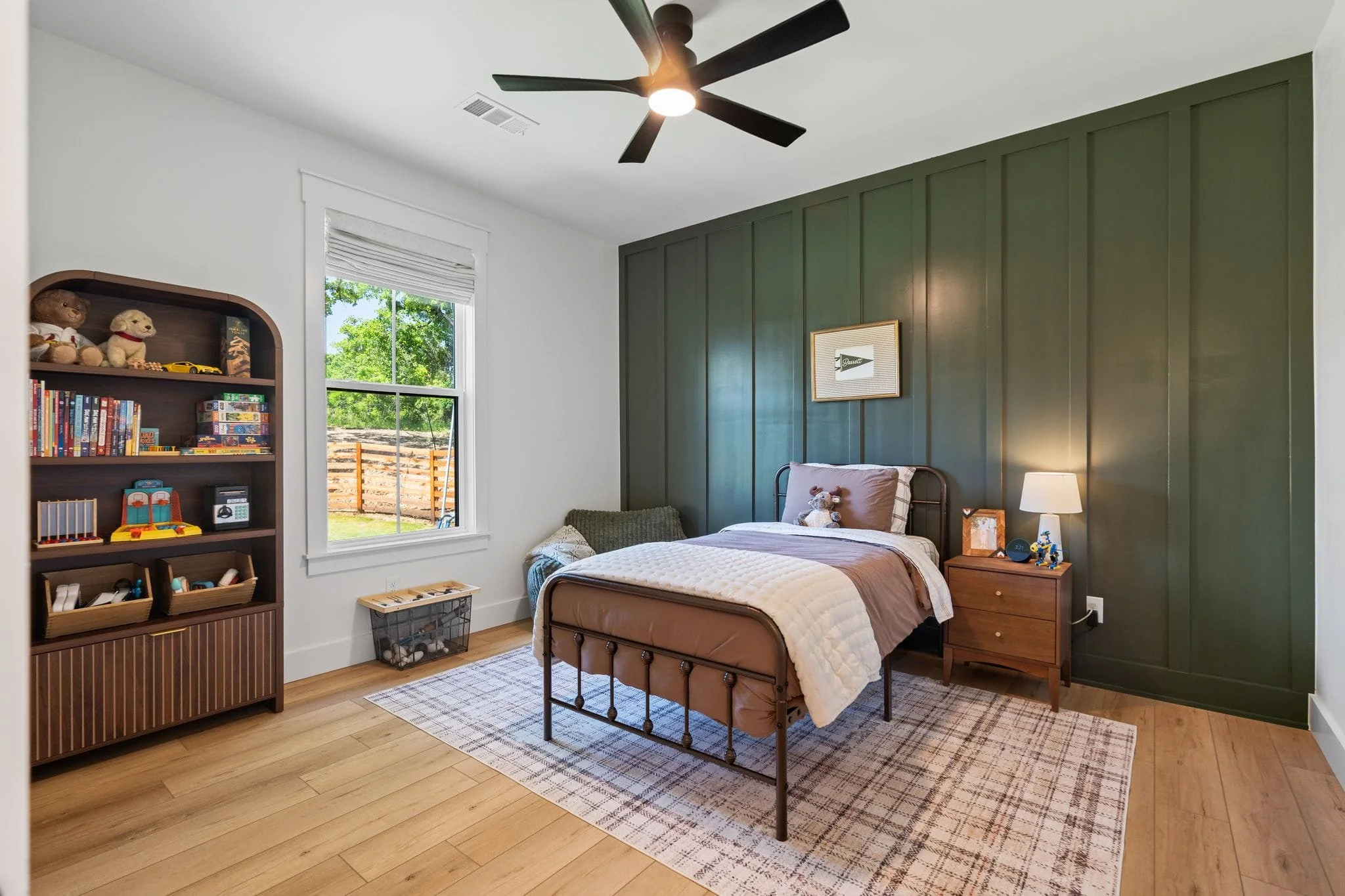 Children’s bedroom with dark green accent wall, metal bed frame, ceiling fan, and wooden bookshelf.