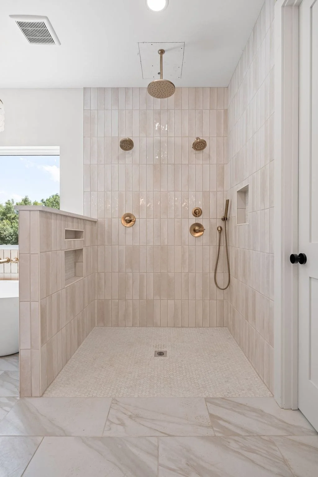 A modern walk-in shower with white tiled walls, three ceiling-mounted shower heads, a handheld shower, and bronze fixtures. There is a small recessed shelf on the wall and a built-in niche for storage. The shower is adjacent to a glass window and has