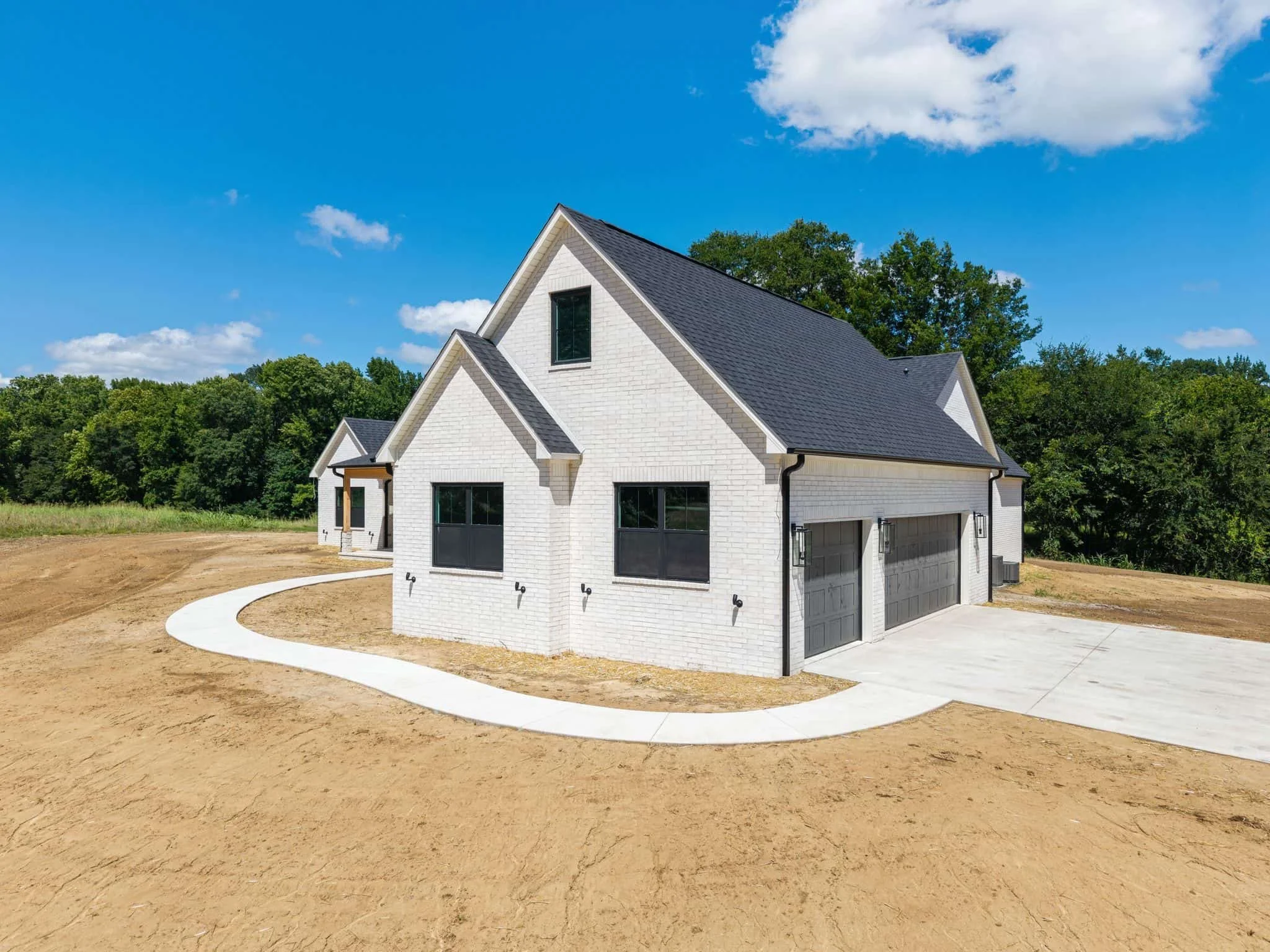 A white house with black roof shingles, large windows, and a garage door, situated on a plot of land with bare soil and a curved concrete pathway, surrounded by green trees under a blue sky with a few clouds.