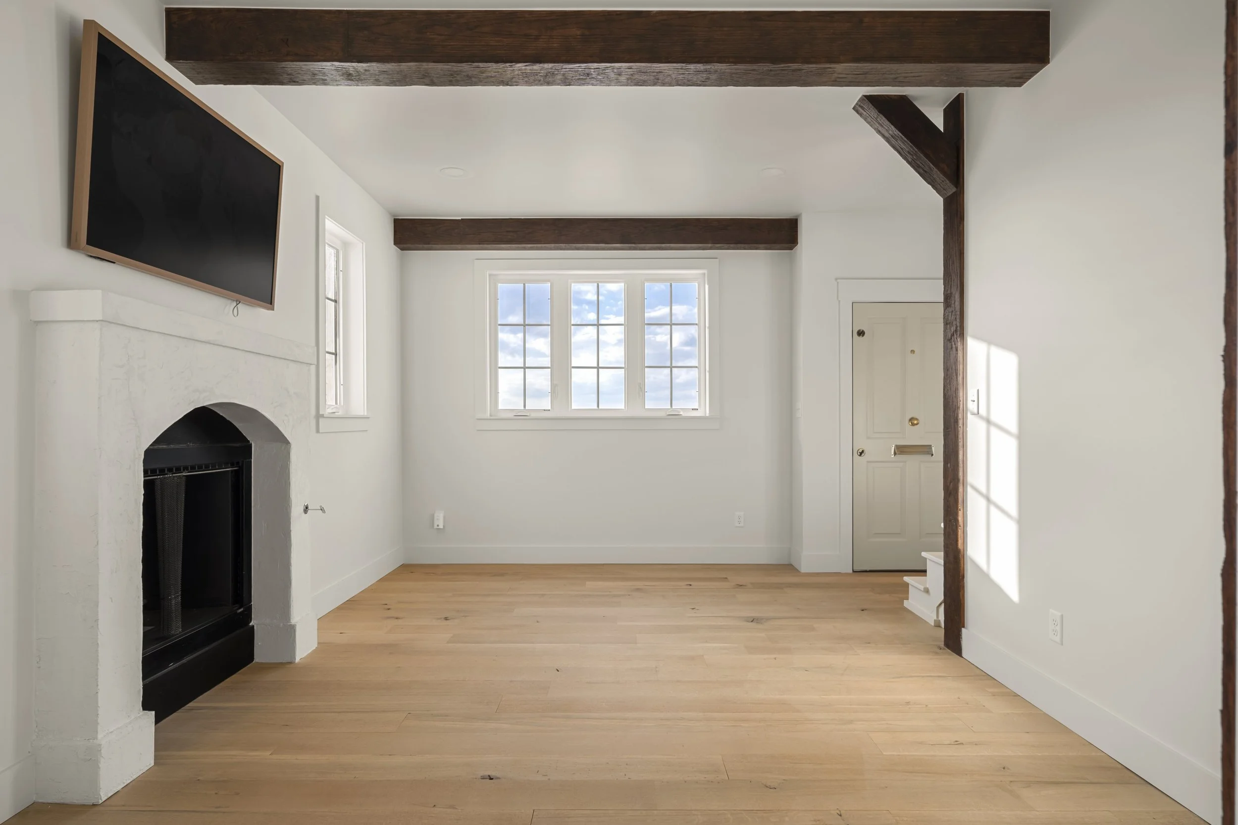 Empty living room with white walls, wooden beams on the ceiling, a large window, a fireplace, and a door, with natural light coming in.