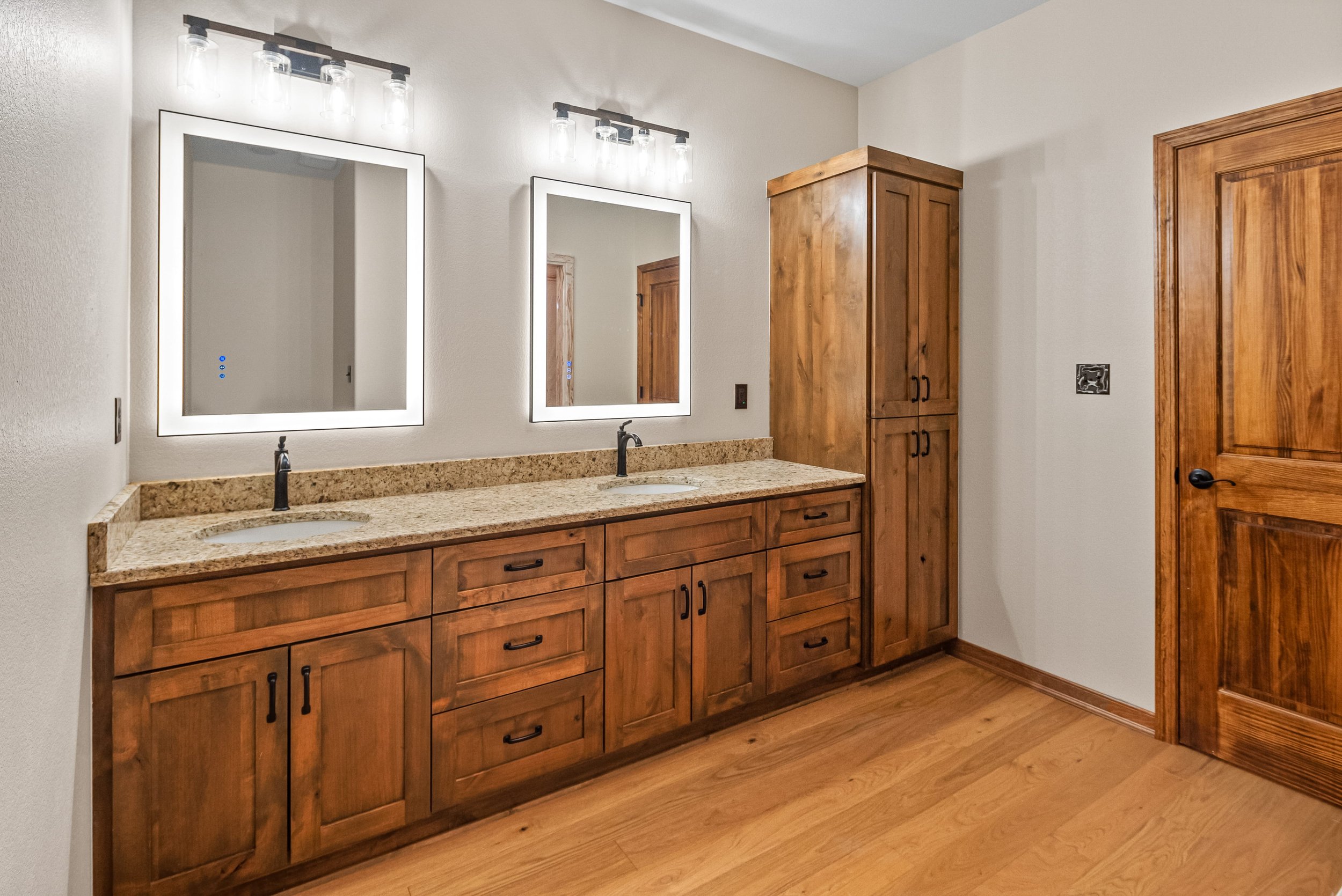 Bathroom with double vanity, granite countertop, two mirrors with backlit frames, wooden cabinetry, and a wooden door.