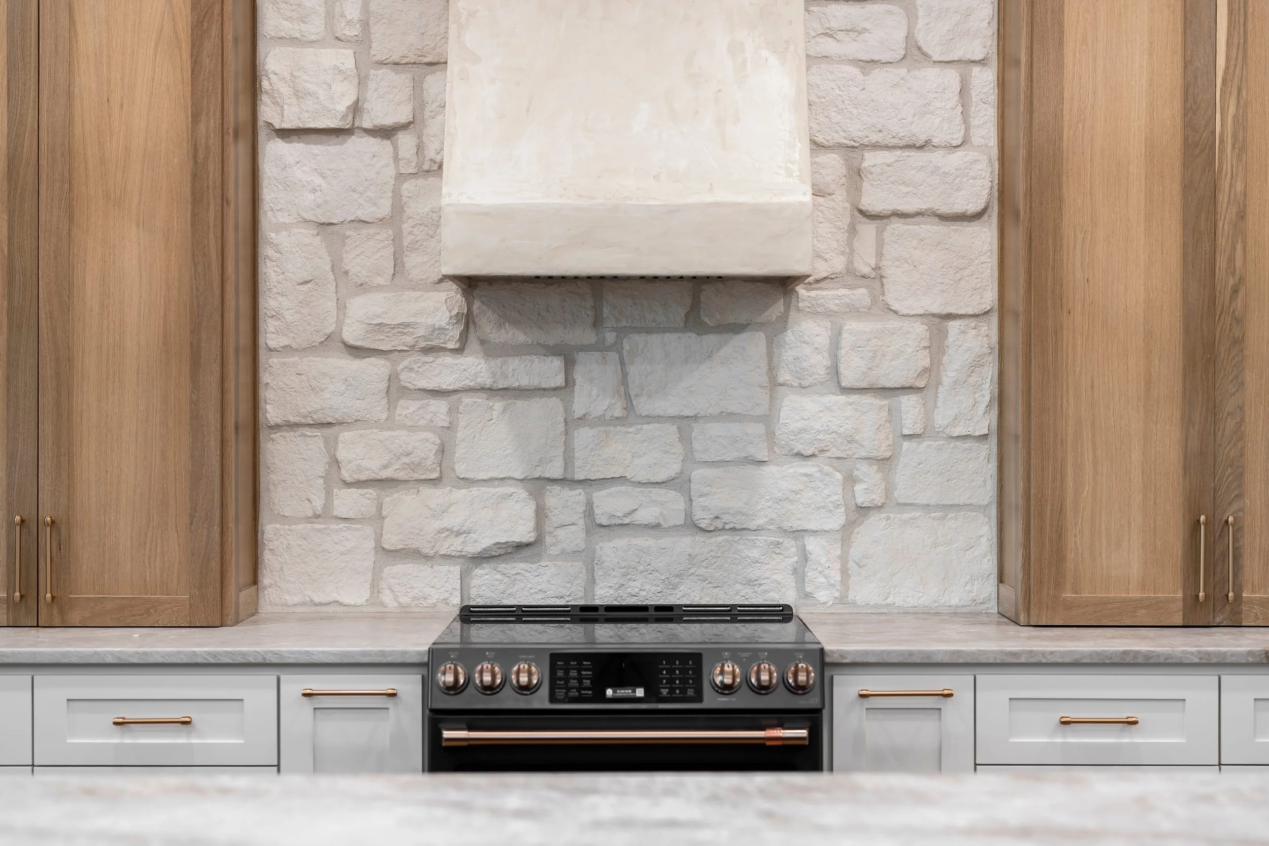 Modern kitchen with white countertops, wooden cabinets, and a black stove with rose gold knobs against a white brick wall.