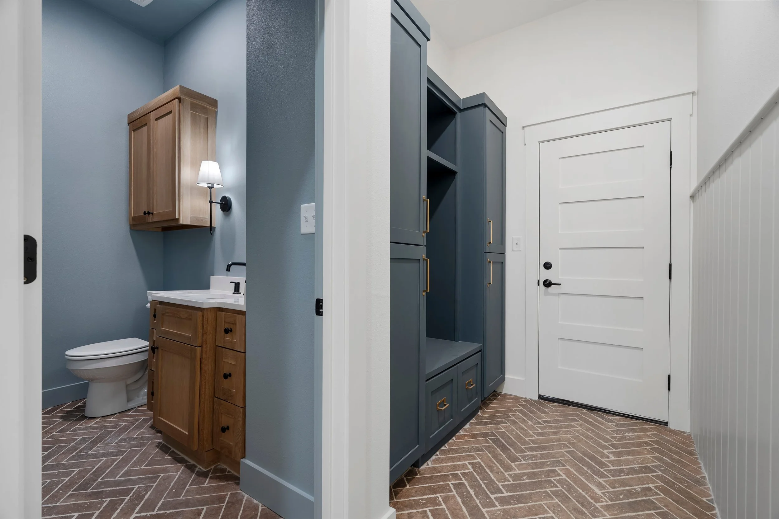 Interior view of a house showing a small bathroom with a wooden cabinet, toilet, and blue walls, and a hallway with a tall blue cabinet, white door, and herringbone brick floor.