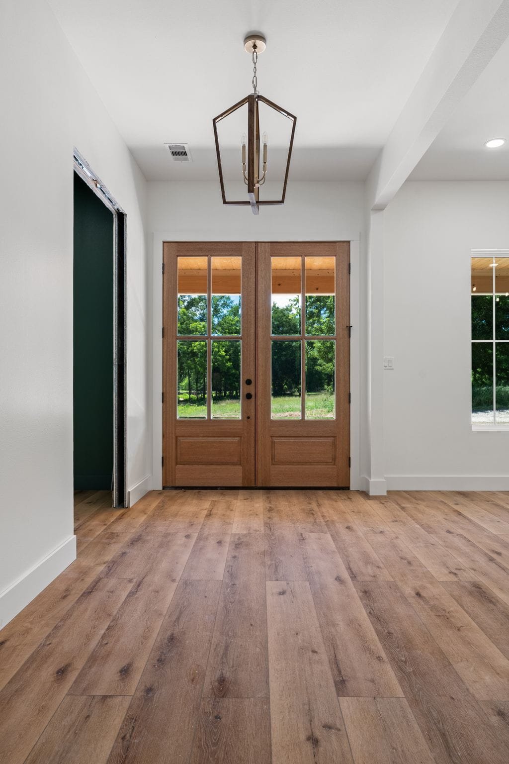 Interior entryway with wooden double doors and large windows showing greenery outside, white walls, hardwood flooring, and a modern chandelier hanging from the ceiling.