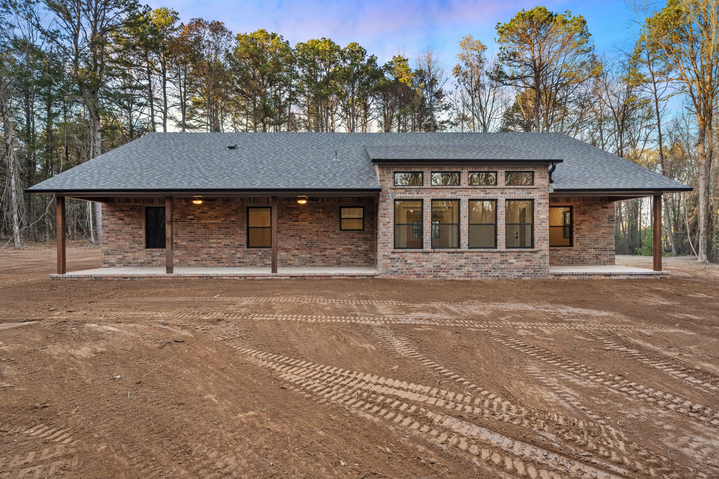 A new brick house with a gray shingled roof, large windows, and a covered porch, set against a wooded background with dirt ground in the foreground.