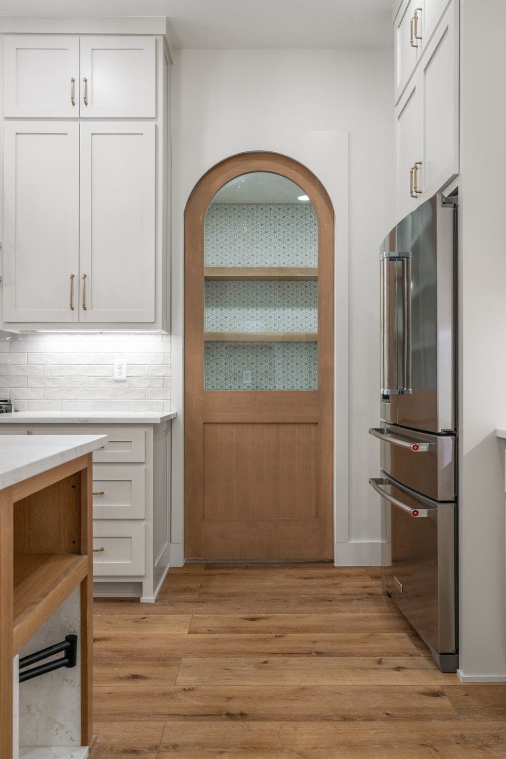 Kitchen with white cabinets, stainless steel refrigerator, wooden floor, and a decorative arched door with patterned wallpaper behind it.