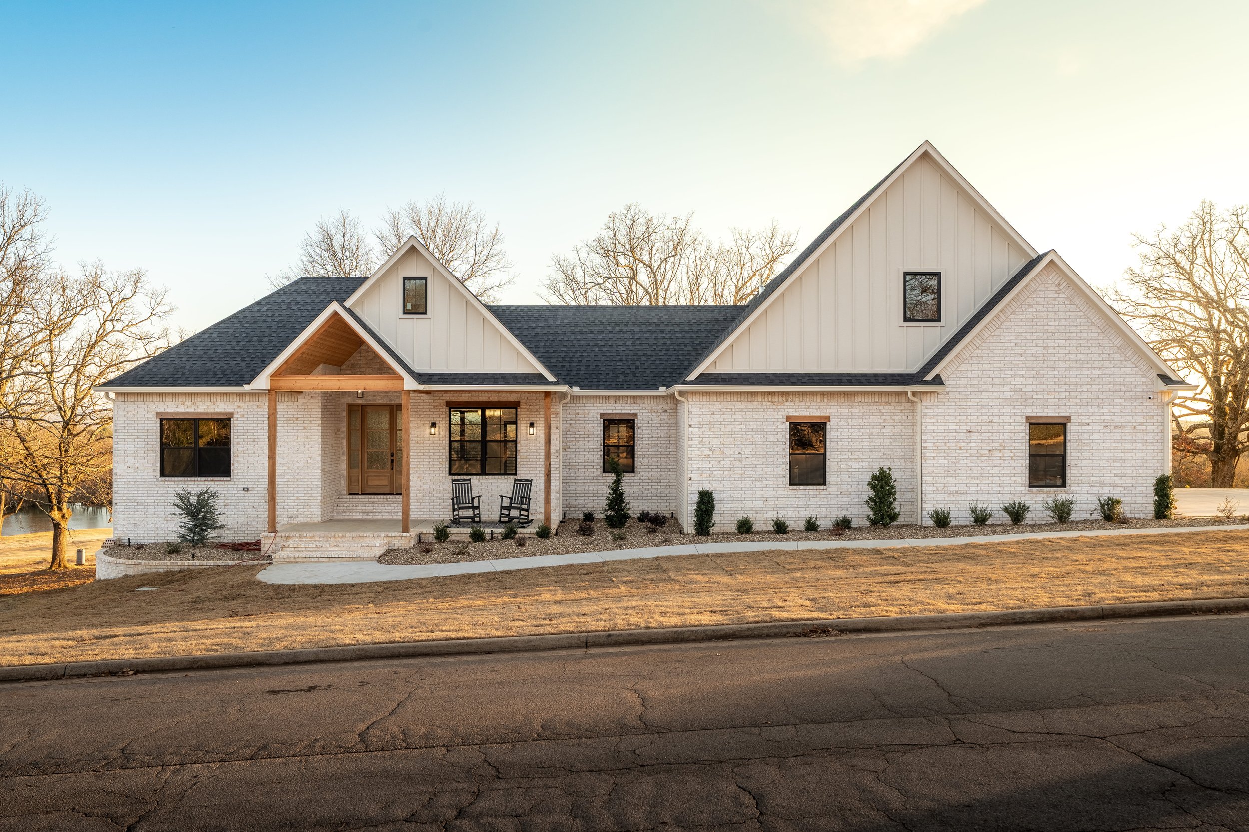 Modern two-story house with white brick exterior, black roof, and front porch, surrounded by landscaped yard and trees in the background.