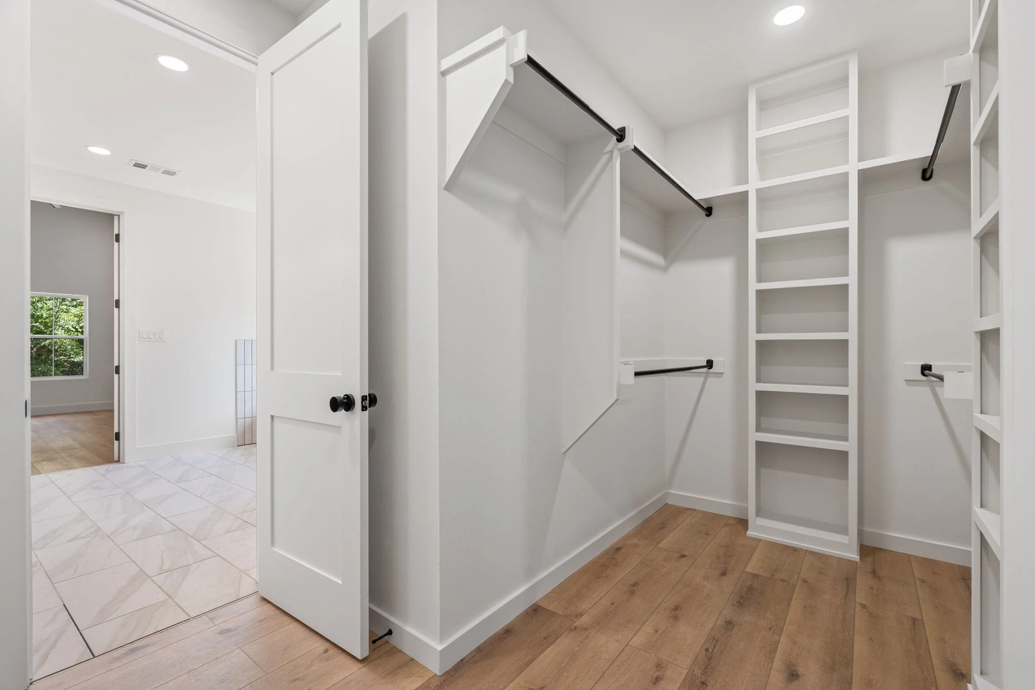 Empty walk-in closet with white shelving, black metal hanging rods, and wooden flooring.
