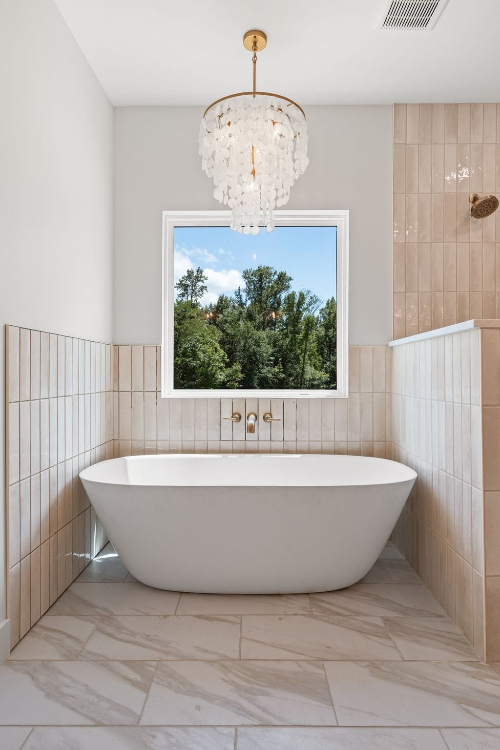 Modern bathroom with a freestanding bathtub below a large window showing trees and a blue sky, with beige tiles and a chandelier.