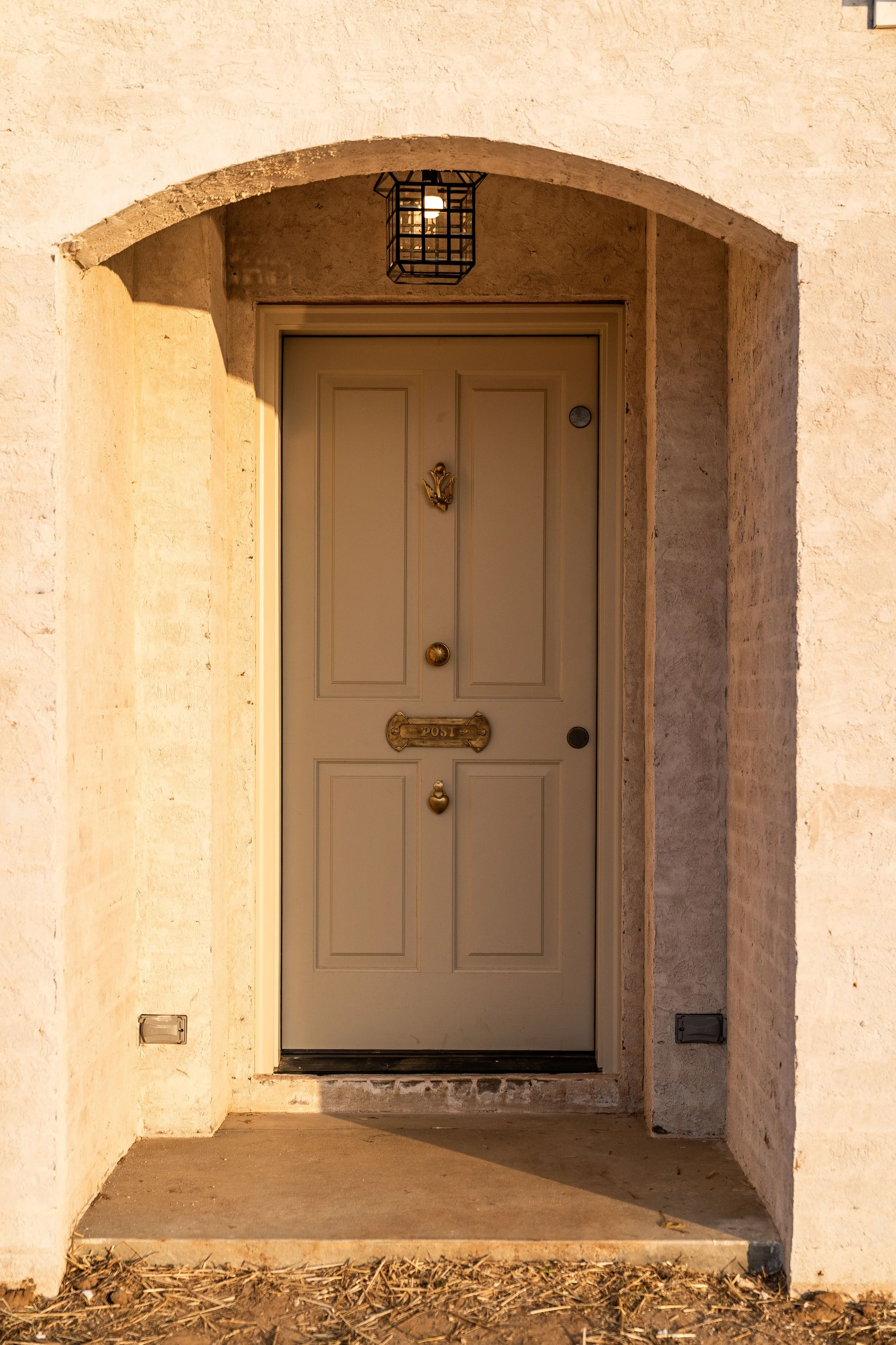 Front door with decorative brass mailbox and door knocker, framed by an arched entrance with a black lantern overhead, adjacent to beige stucco walls.