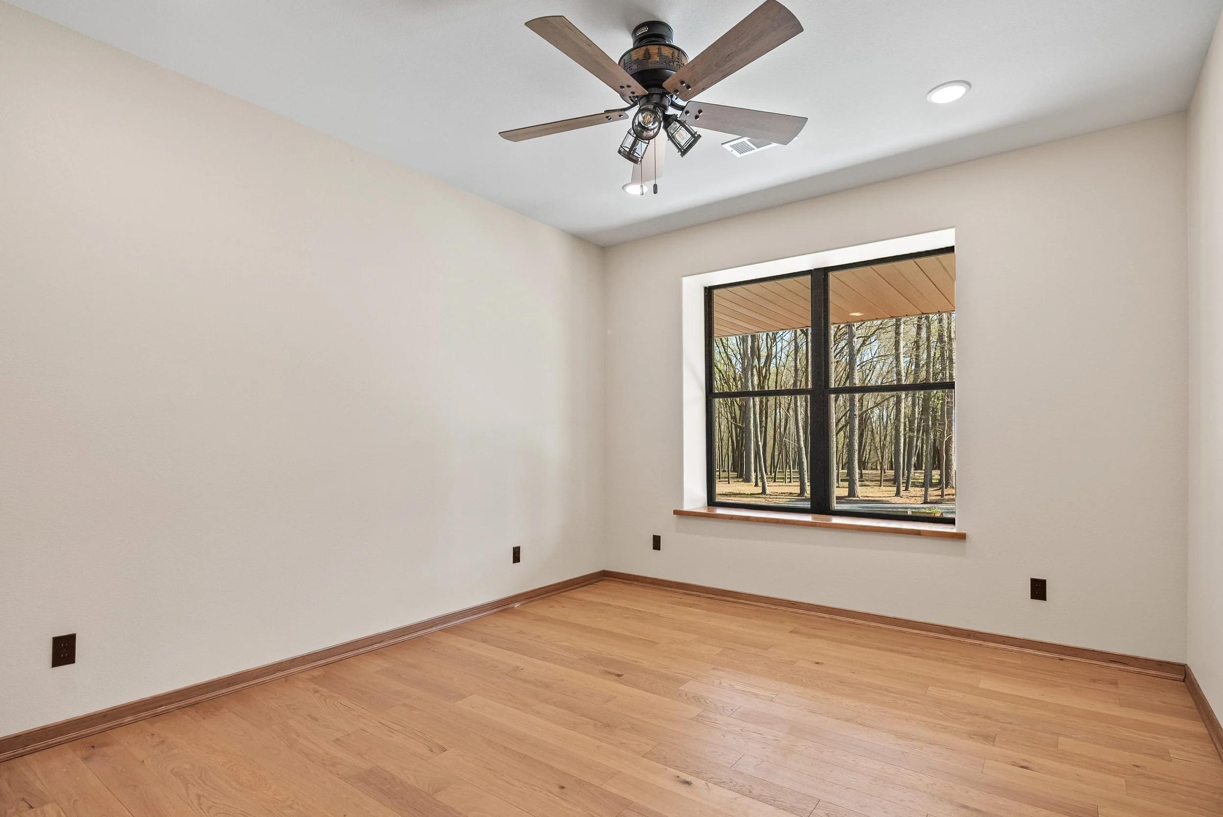 Empty room with beige walls, hardwood flooring, a large window showing trees outside, and a ceiling fan with lights.