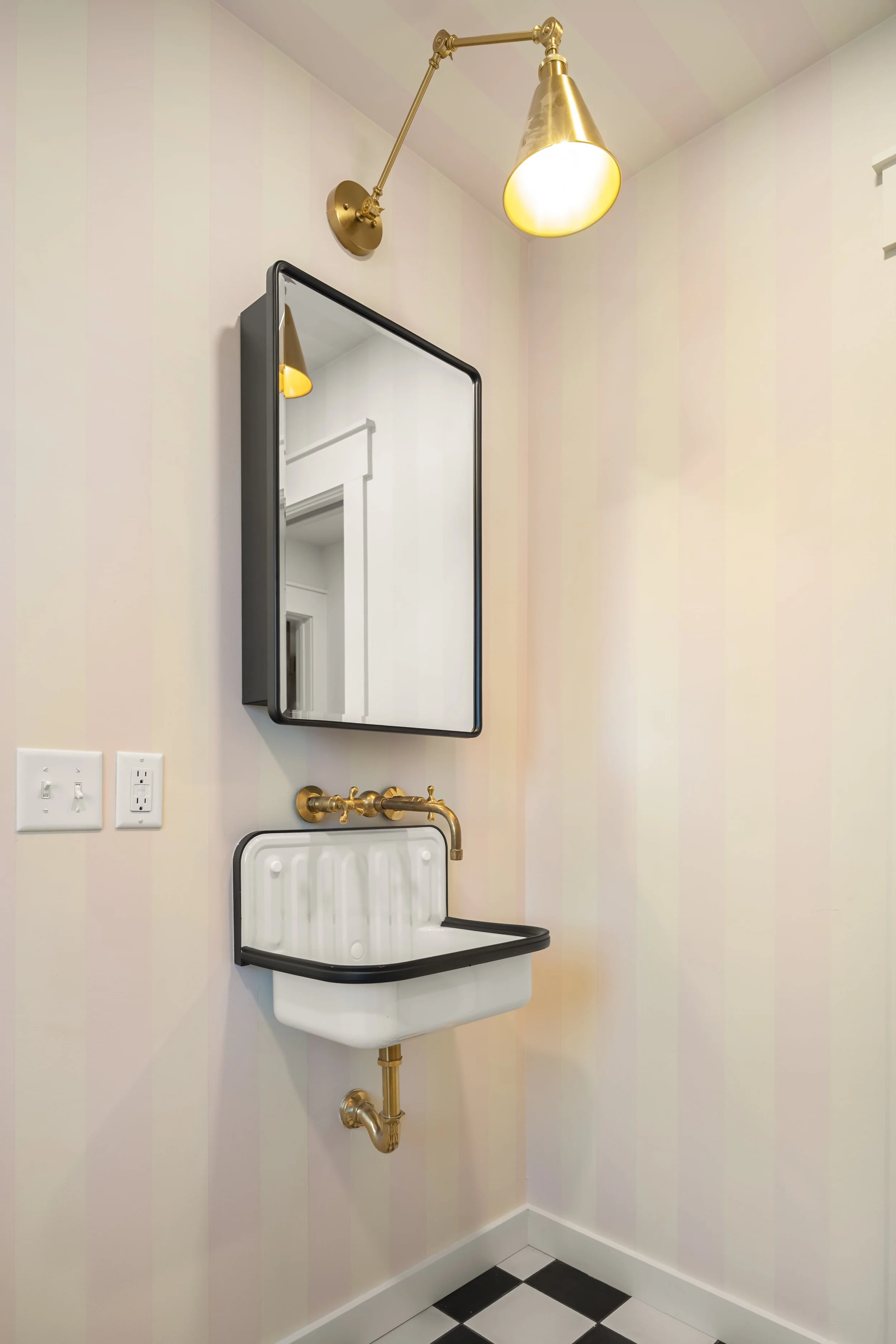 Small wall-mounted sink with gold faucet, mirror above it, wall sconce light fixture, and electrical outlets on a striped wall in a bathroom.