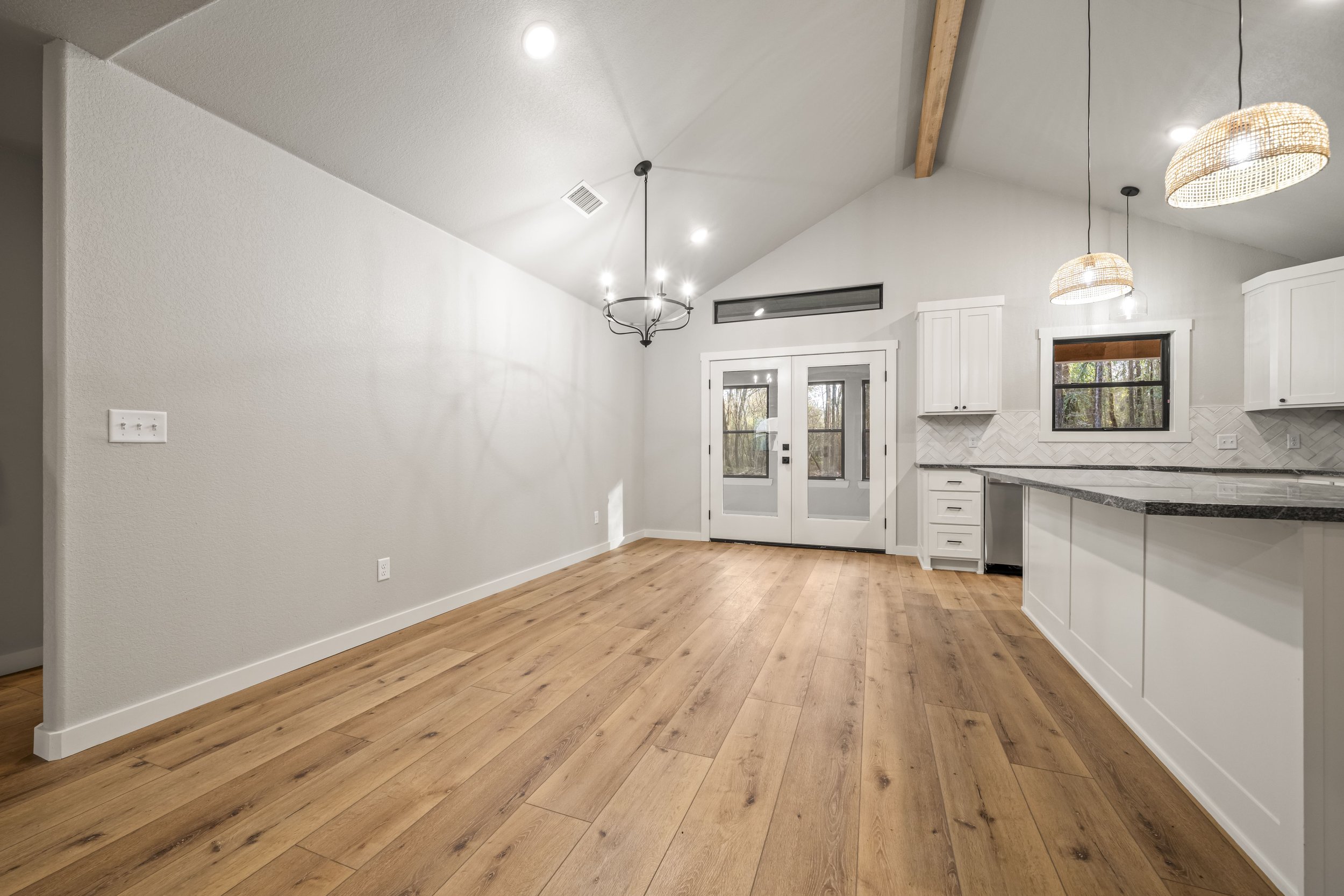 Empty dining area and kitchen with hardwood floors, white walls, white cabinets, pendant lights, French doors, and windows showing a wooded outdoor view.