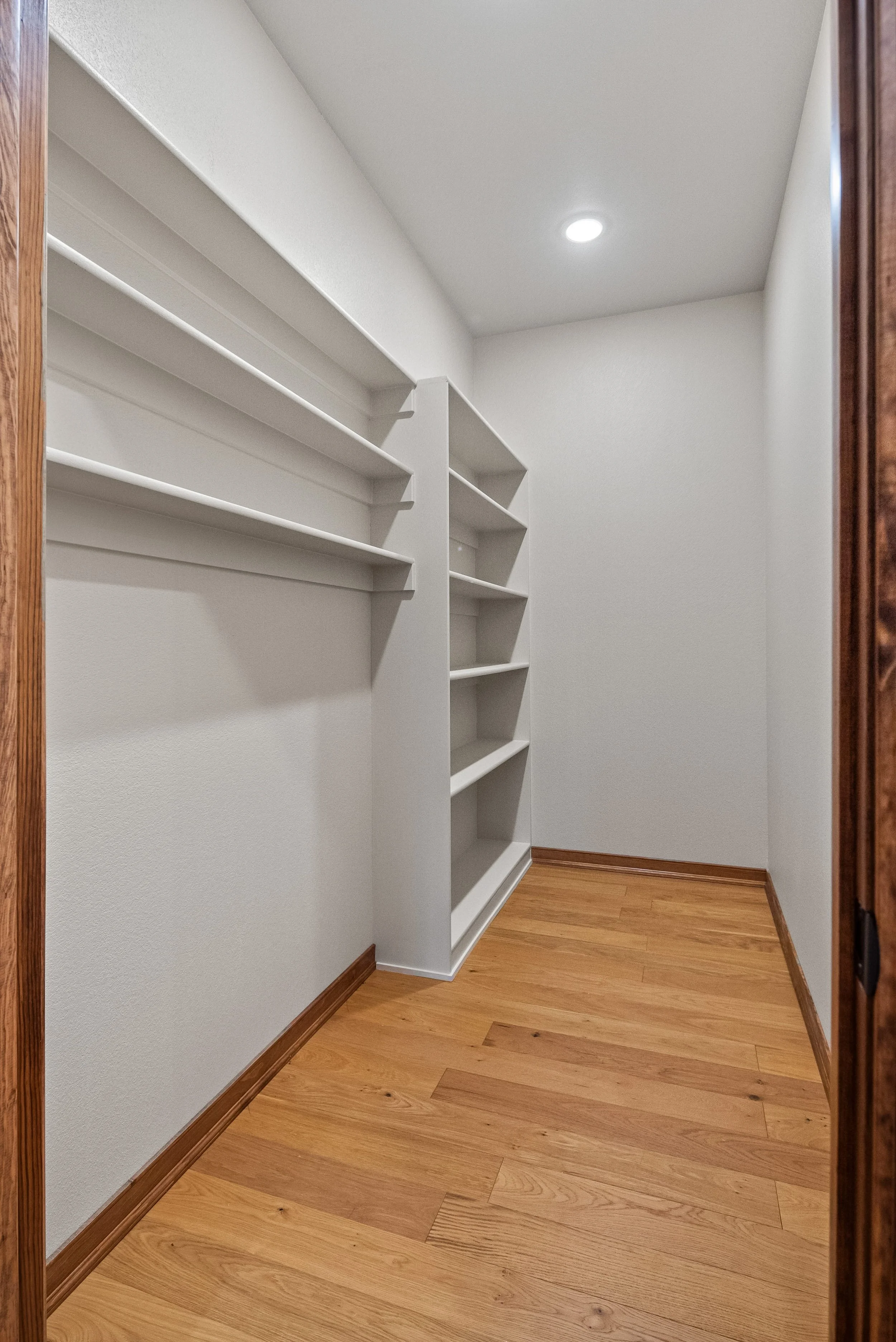 Empty walk-in closet with white shelves and wooden trim, hardwood floor, and ceiling light.