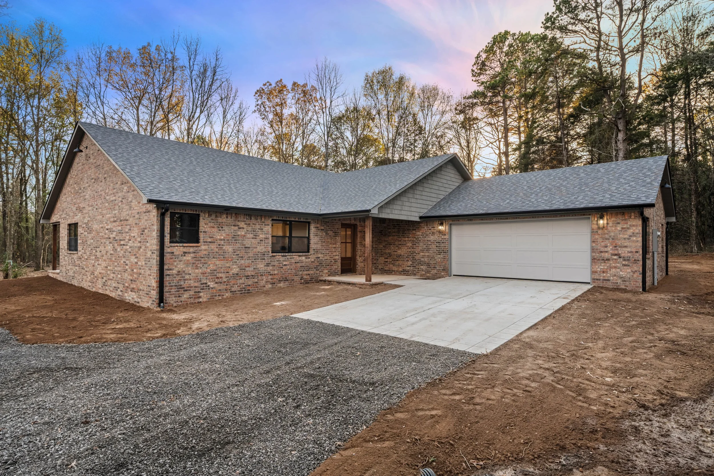Newly built single-story brick house with a gray garage door, a concrete driveway, and a small front porch, surrounded by dirt and bare trees under a sunset sky.