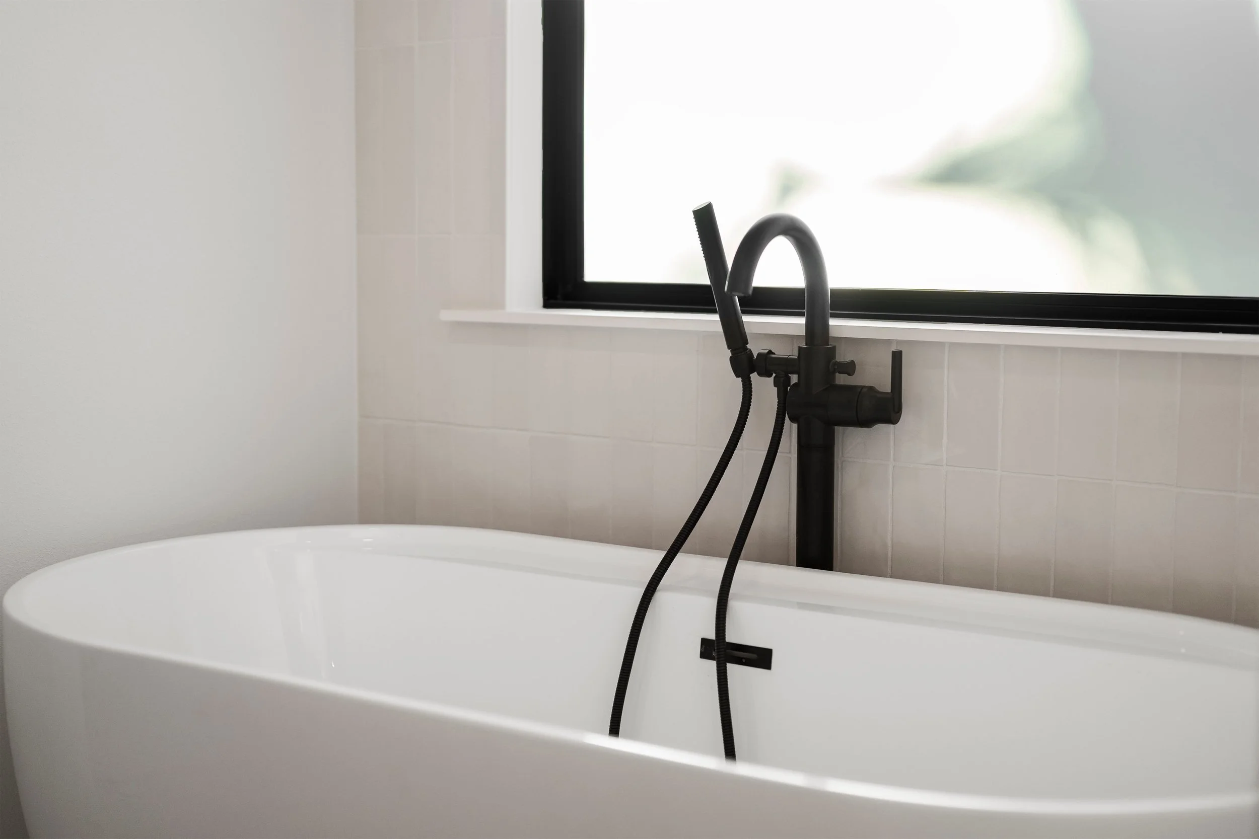 White freestanding bathtub with black faucet and handheld shower near a window.