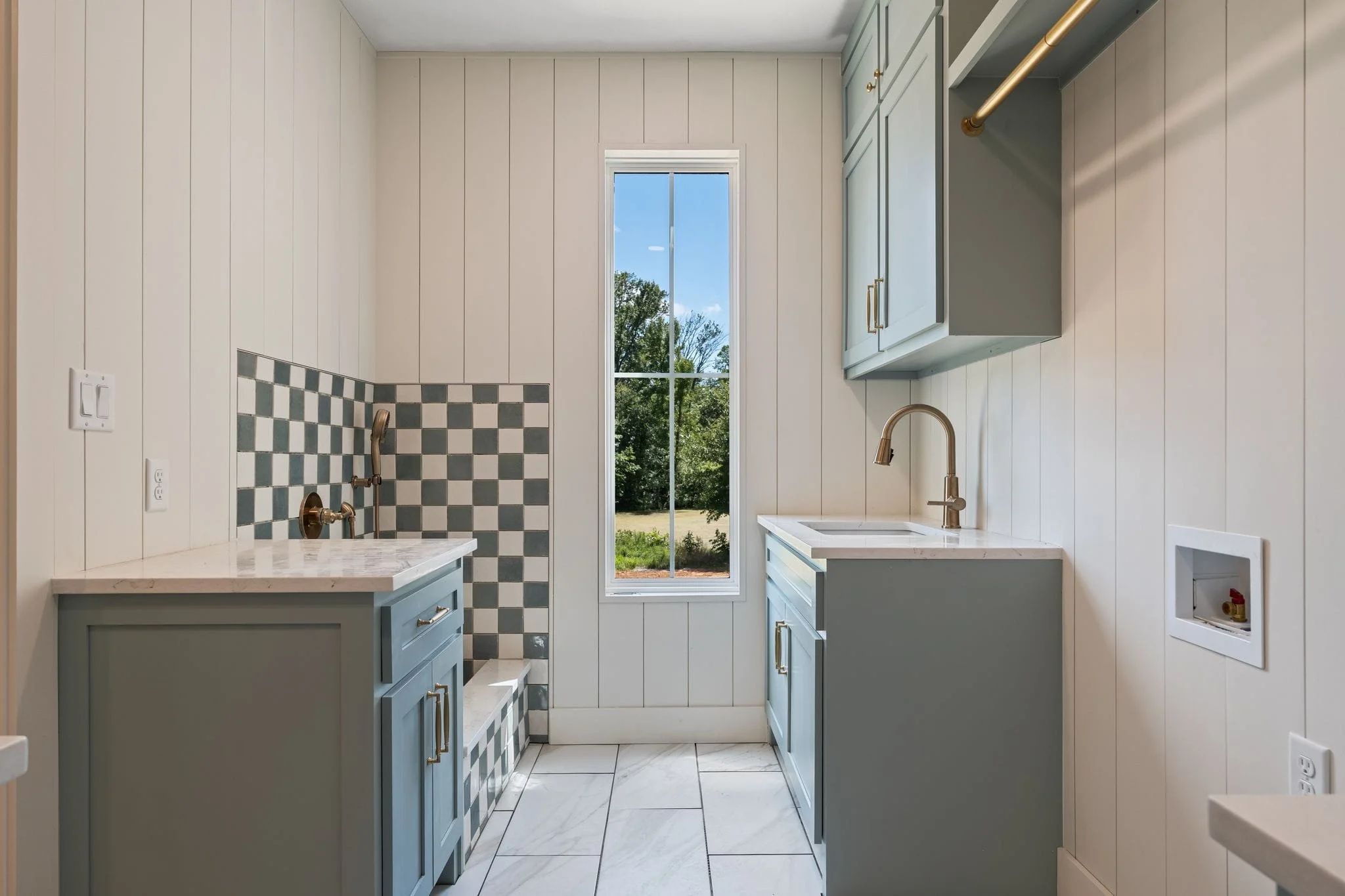 A small kitchen with light blue cabinets, white countertops, and a large window showing trees outside. There are copper fixtures and a checkered tile wall in the corner.