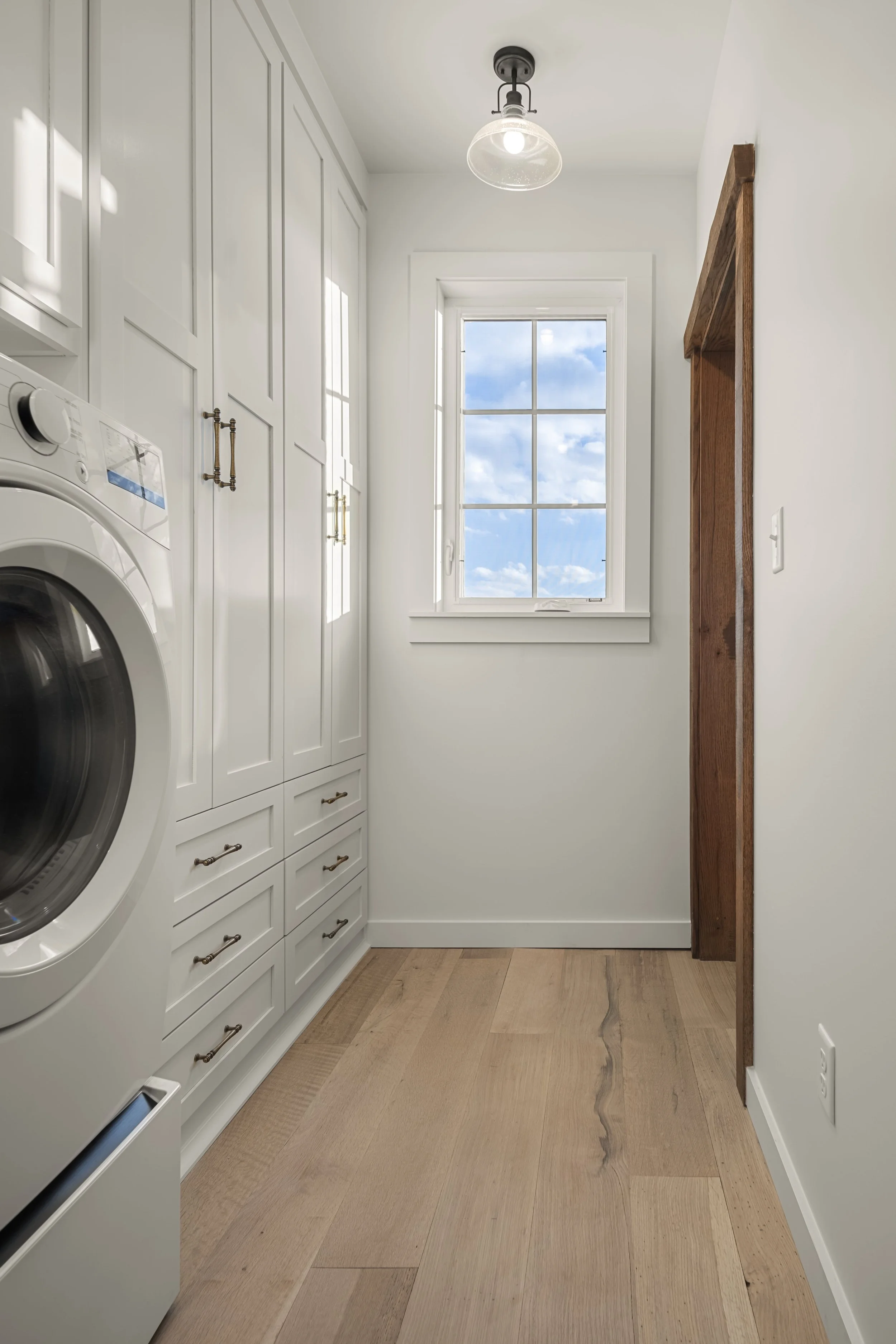 A laundry room with white cabinetry, a washing machine, a window showing a blue sky with clouds, and wooden accents.