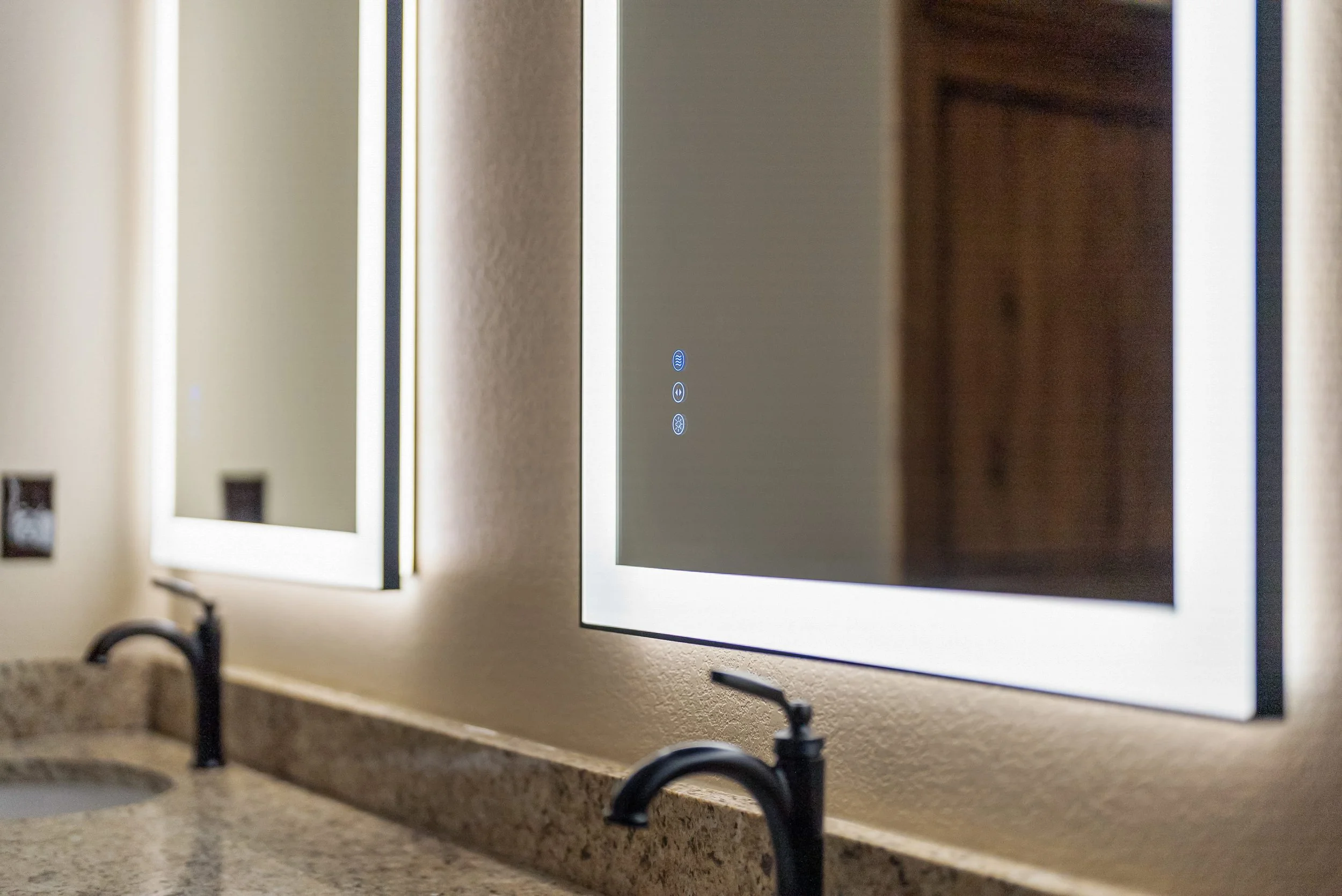 Close-up view of two illuminated mirrors with embedded digital control panels on a bathroom wall above a granite sink with black faucets.