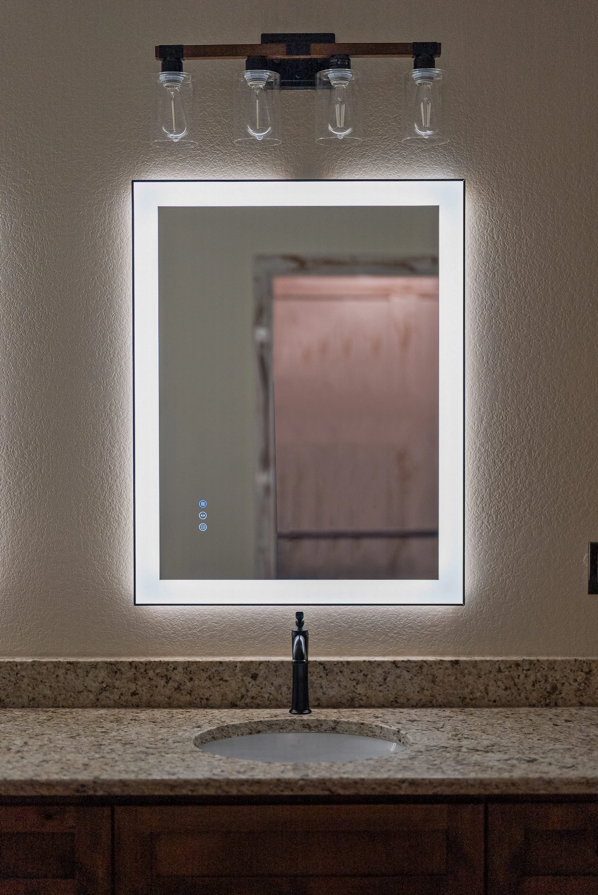 Modern bathroom mirror with LED lighting, mounted above a granite countertop with a sink and black faucet. A decorative light fixture with glass shades hangs above the mirror. The wall behind the mirror is beige.