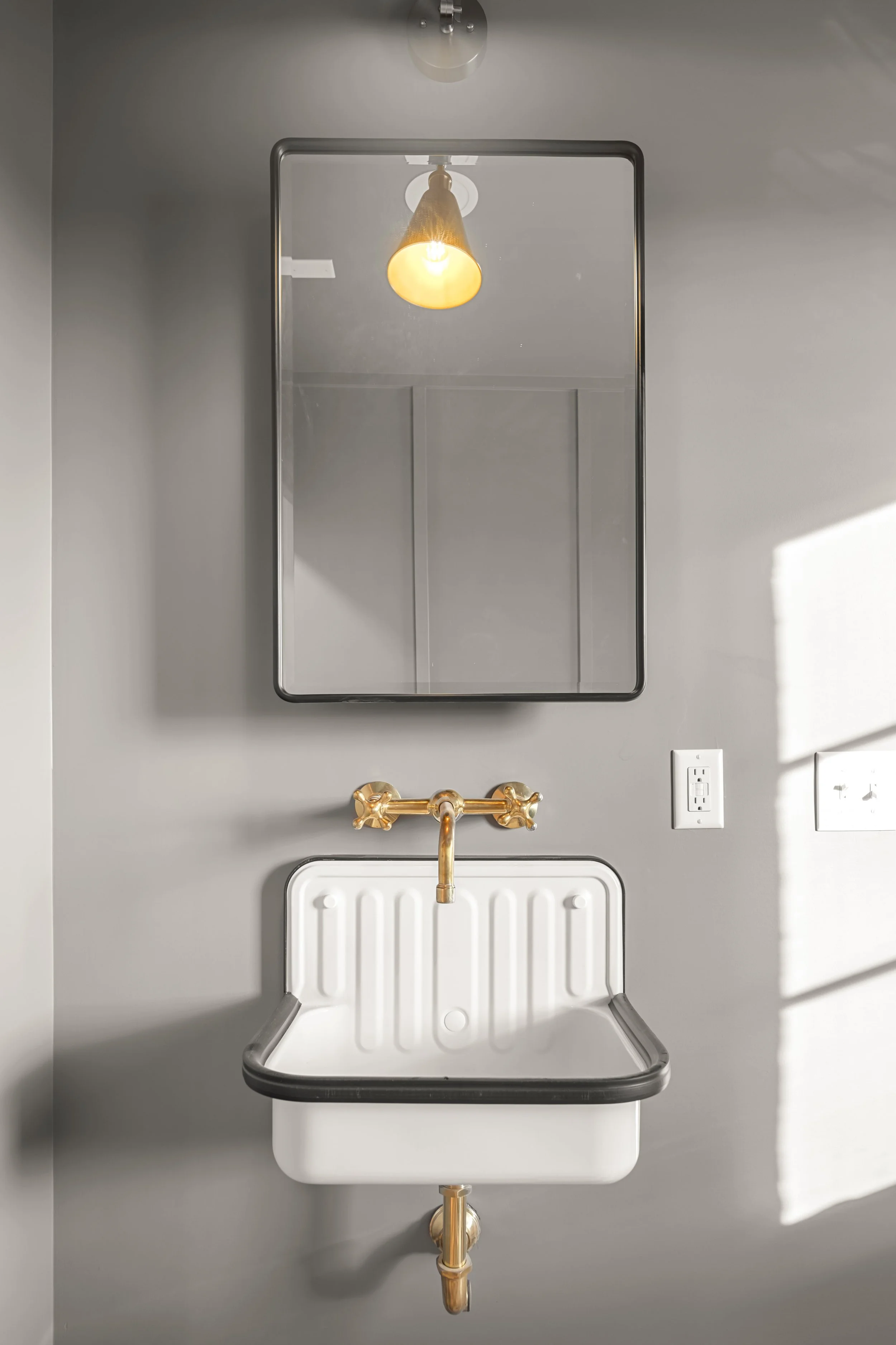 Small white wall-mounted sink with black trim, gold faucet, rectangular mirror above, gray wall, electrical outlet on the right, and sunlight casting shadows on the wall.