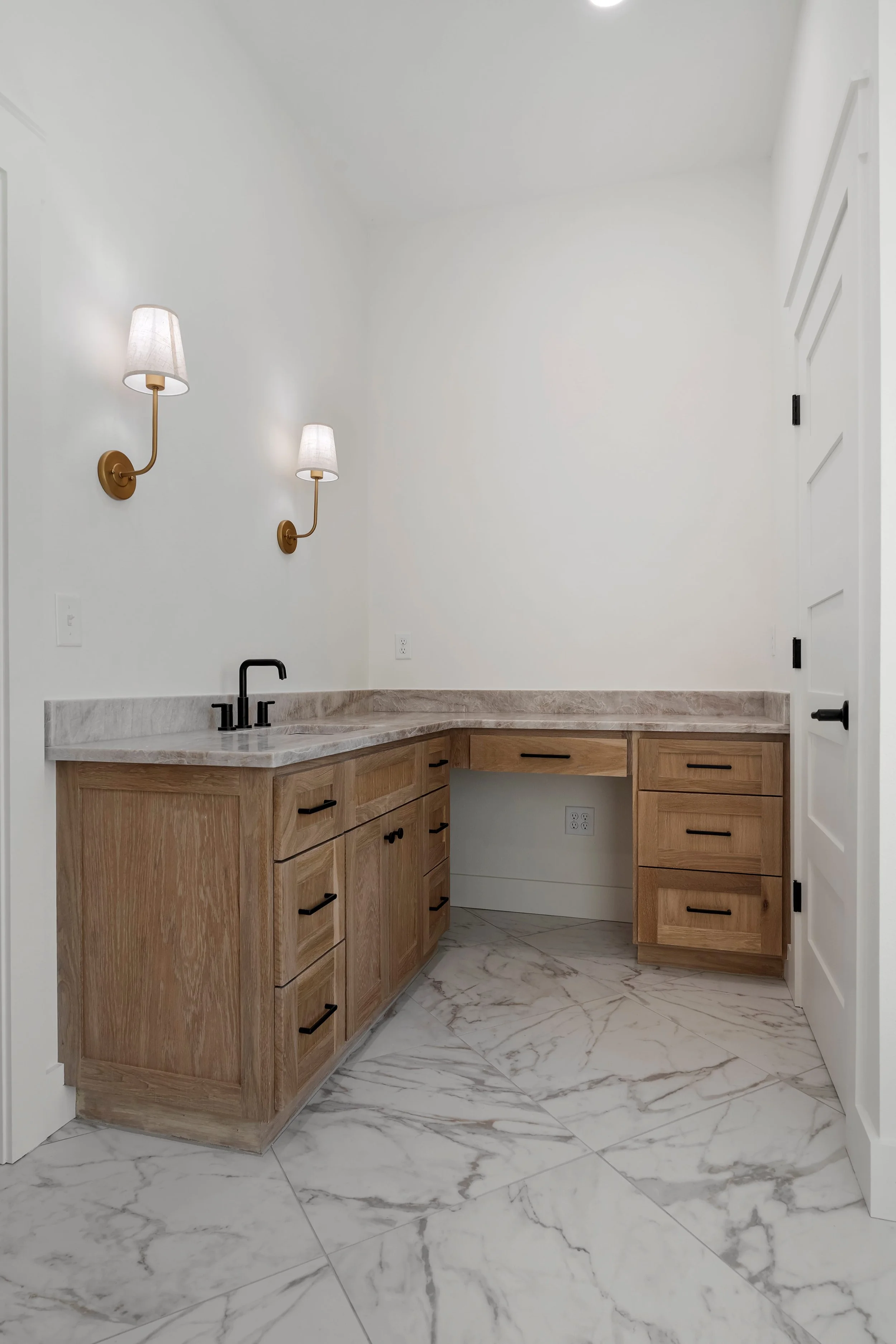 A room with a marble floor, wooden cabinets, a countertop, two wall-mounted lamps, and a partially open door.