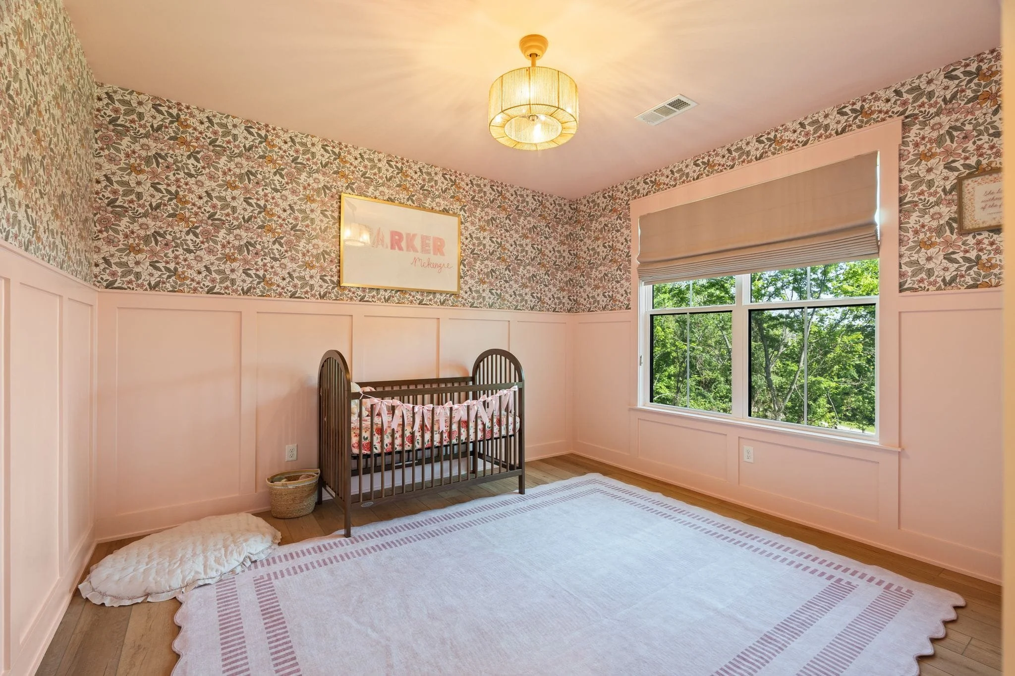 Pink nursery with floral wallpaper, wood crib, wainscoting, and large window with tree views.
