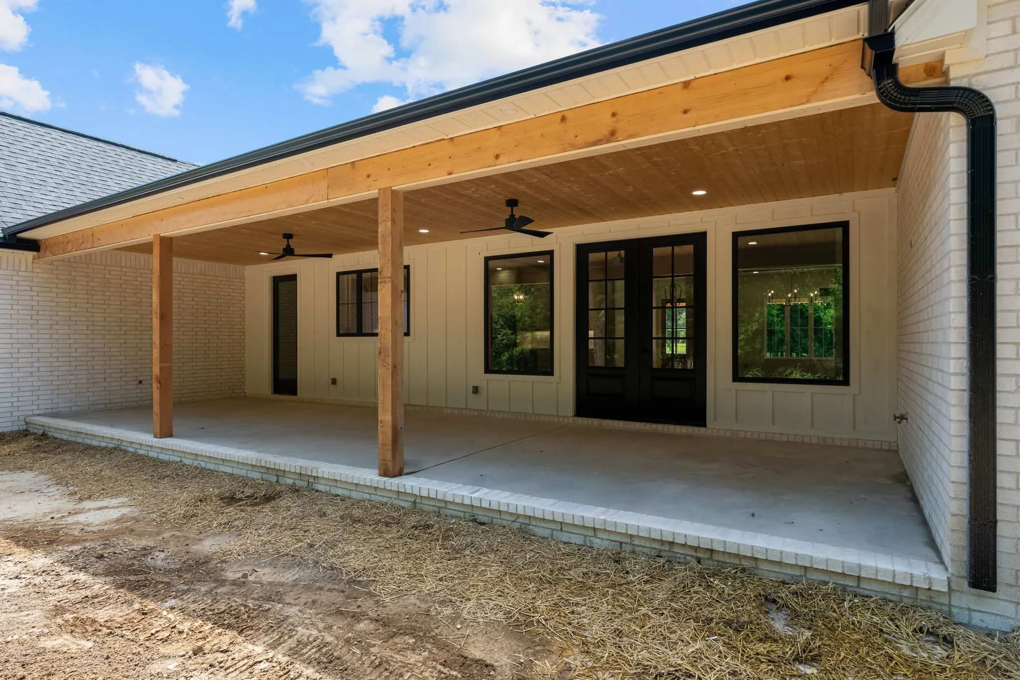 A covered patio area at the back of a house with a ceiling fan, recessed lighting, and large glass doors and windows, with a dirt and straw ground surface.