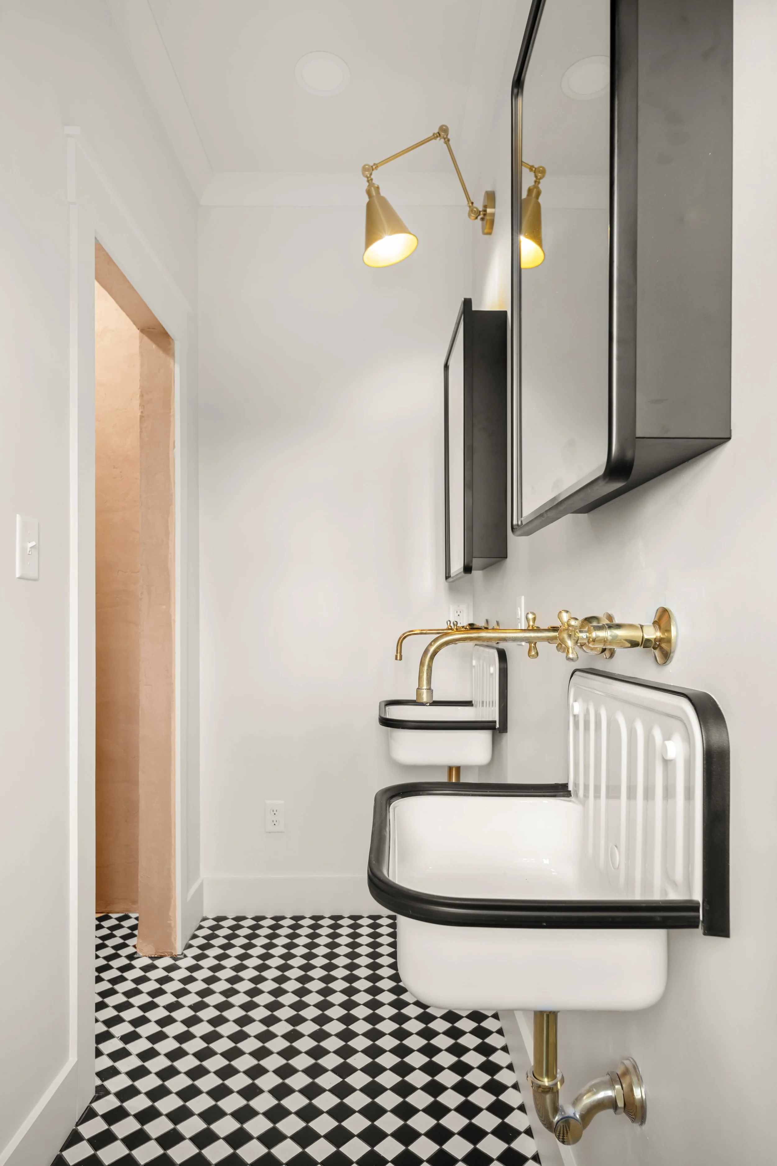 A small bathroom with black and white checkered floor, a white wall, a black wall-mounted cabinet, a hanging mirror, a brass wall sconce, a vintage wall-mounted sink with brass faucet, and a black and white wall-mounted laundry tub.