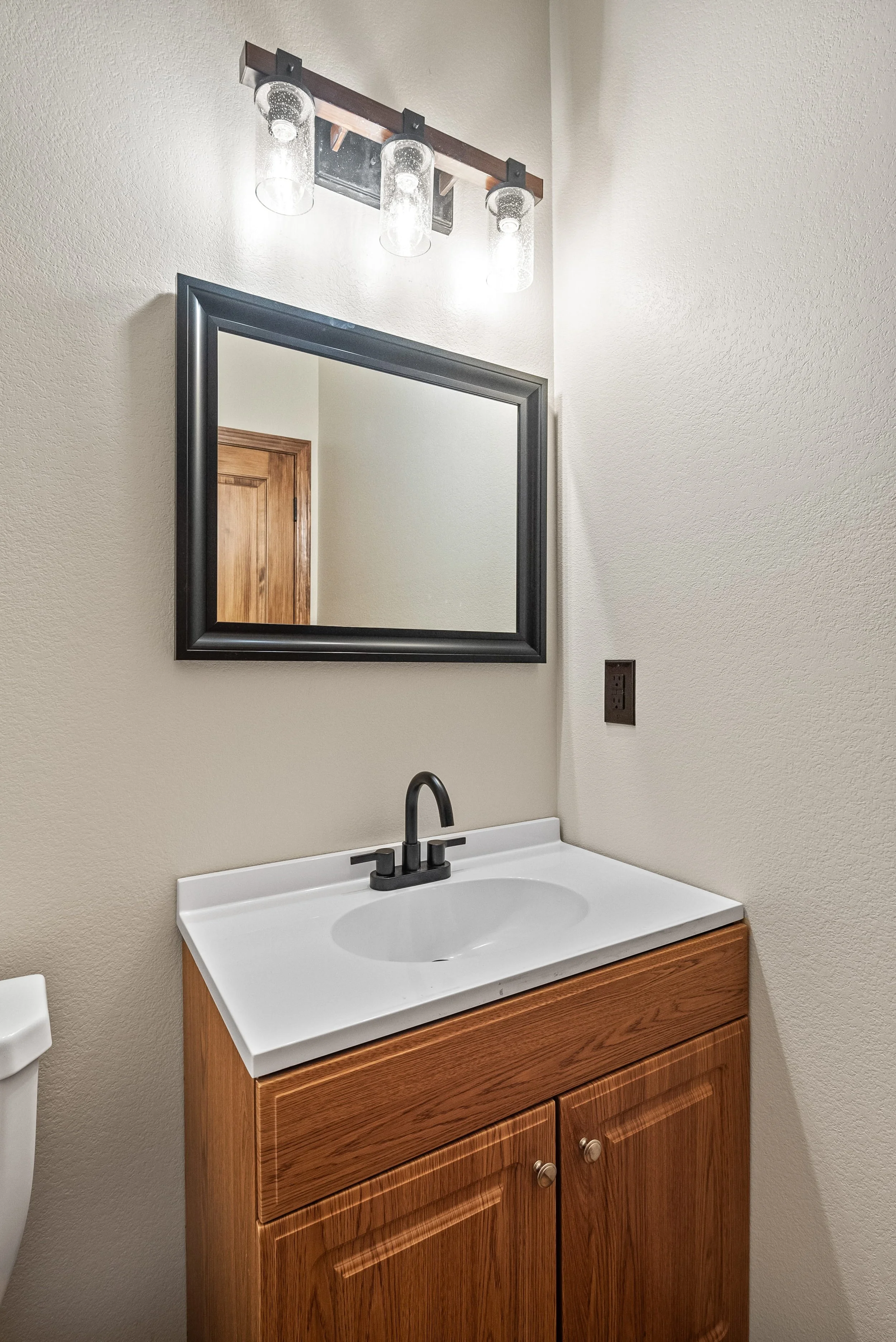 Bathroom sink with a black faucet, a mirror above, and a wooden cabinet below, with a light fixture with three glass shades above.
