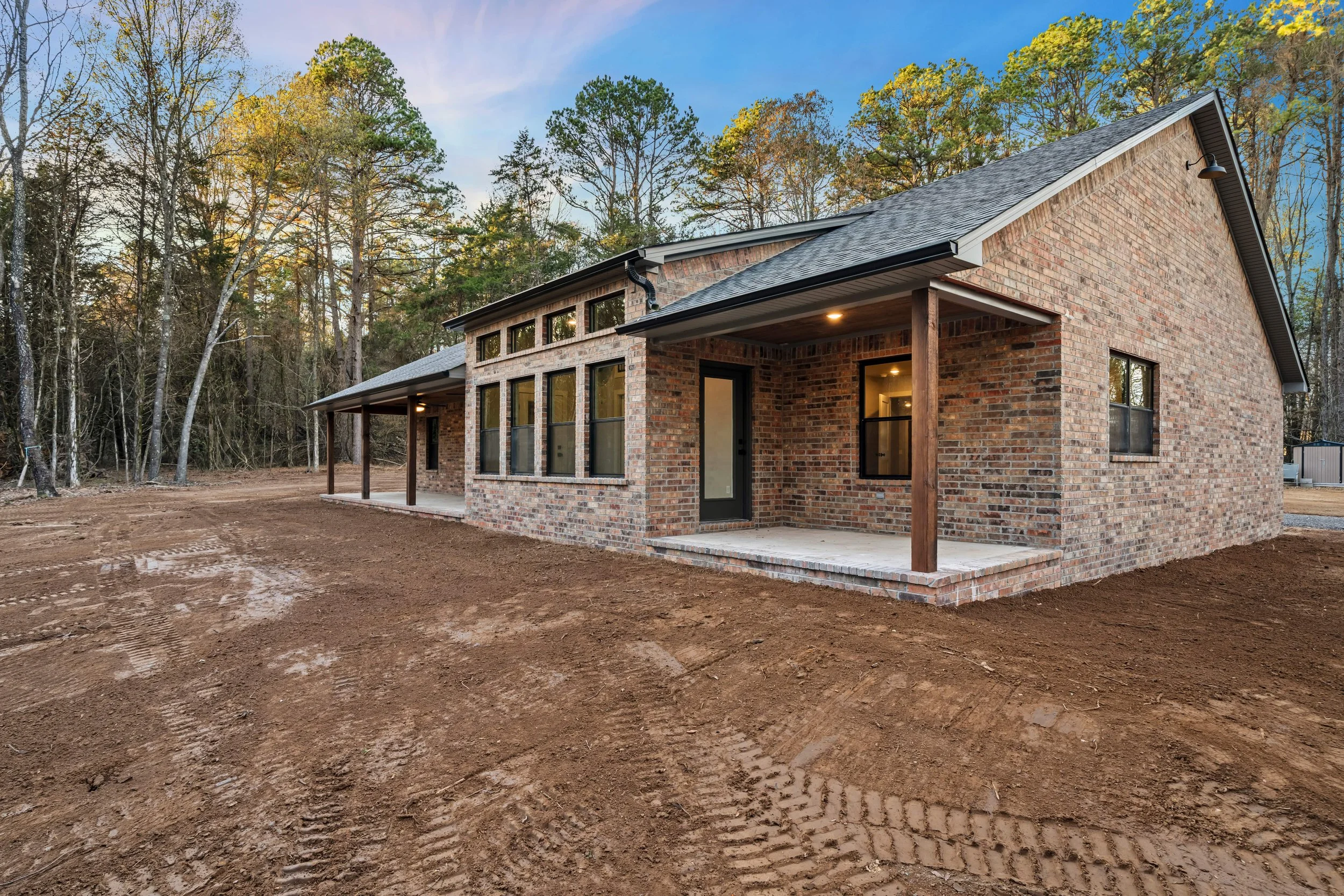 New brick house with large windows and covered porch, set in a wooded area during dusk, with dirt ground in the foreground.