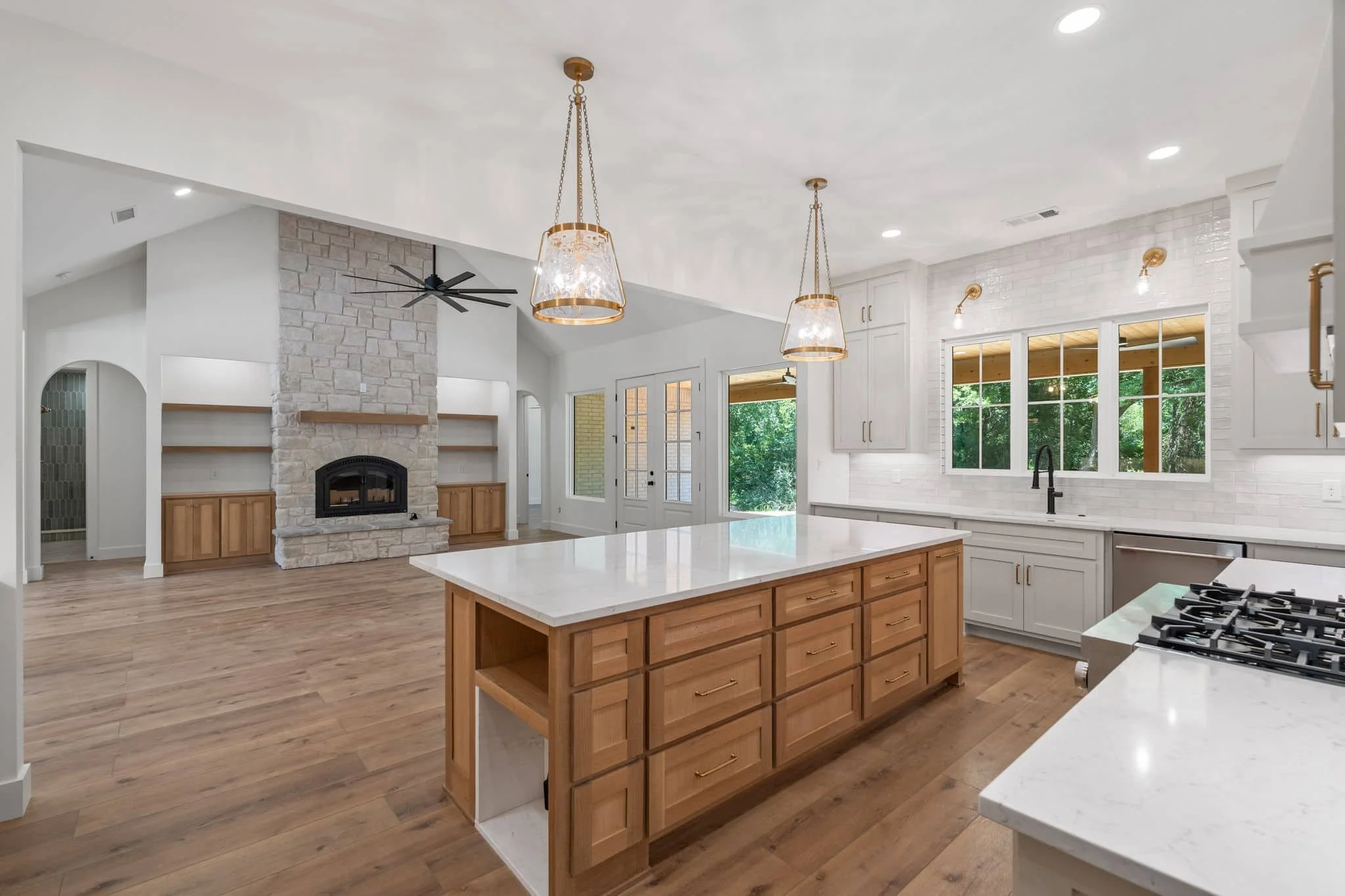 Modern kitchen with white cabinets, a large island with wood drawers, and pendant lights. There is a stone fireplace in the background, large windows showing greenery outside, and a ceiling fan.