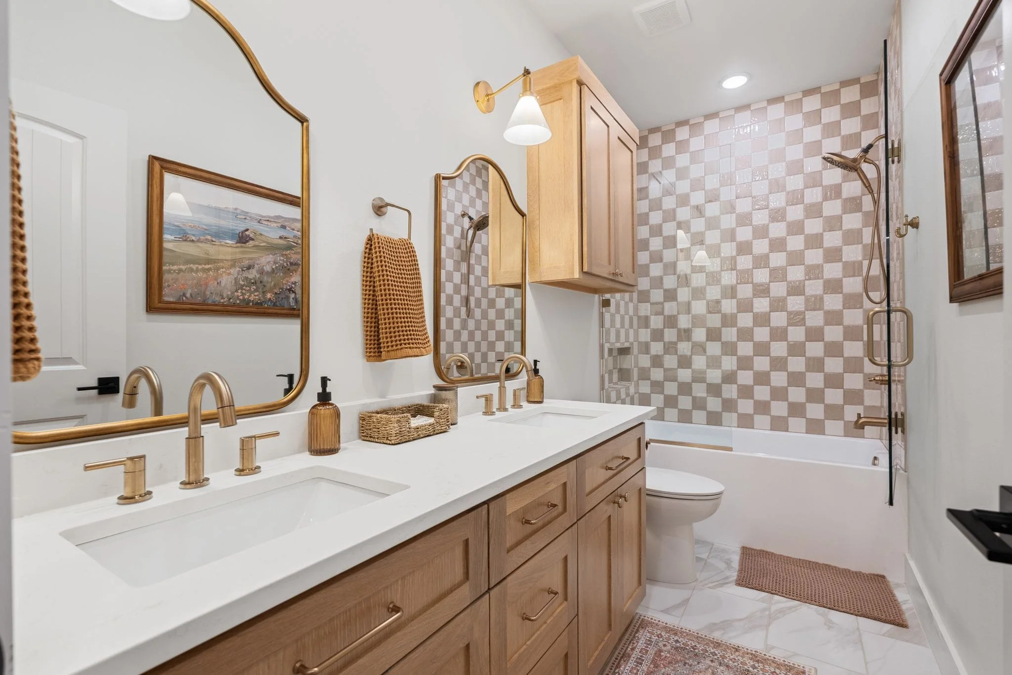 Modern bathroom with double vanity, gold fixtures, checkered tile shower surround, and framed mirrors.
