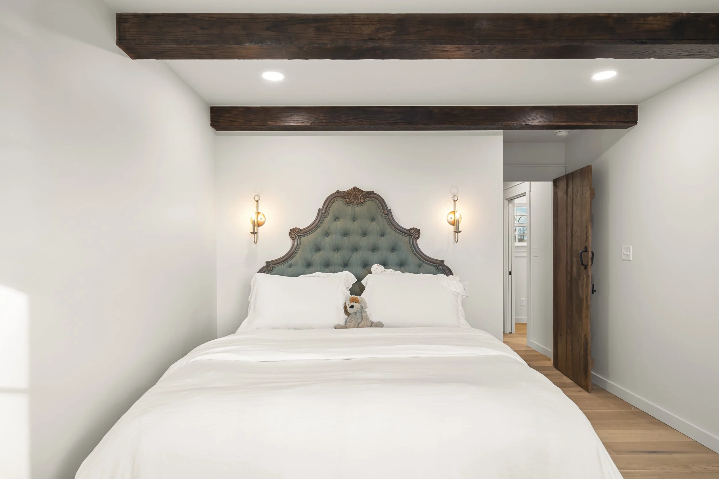 A bedroom with a large bed featuring a tufted pastel blue headboard, white bedding, and a stuffed animal on the pillow. Two wall-mounted lamps flank the headboard, and the room has white walls, wooden beams on the ceiling, and a wooden door.