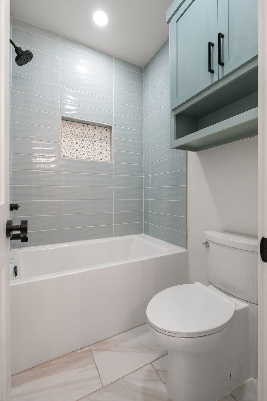 A small modern bathroom with a white bathtub, light blue tiled wall, a built-in niche with decorative tile, a ceiling-mounted showerhead, a toilet, and light-colored floor tiles.