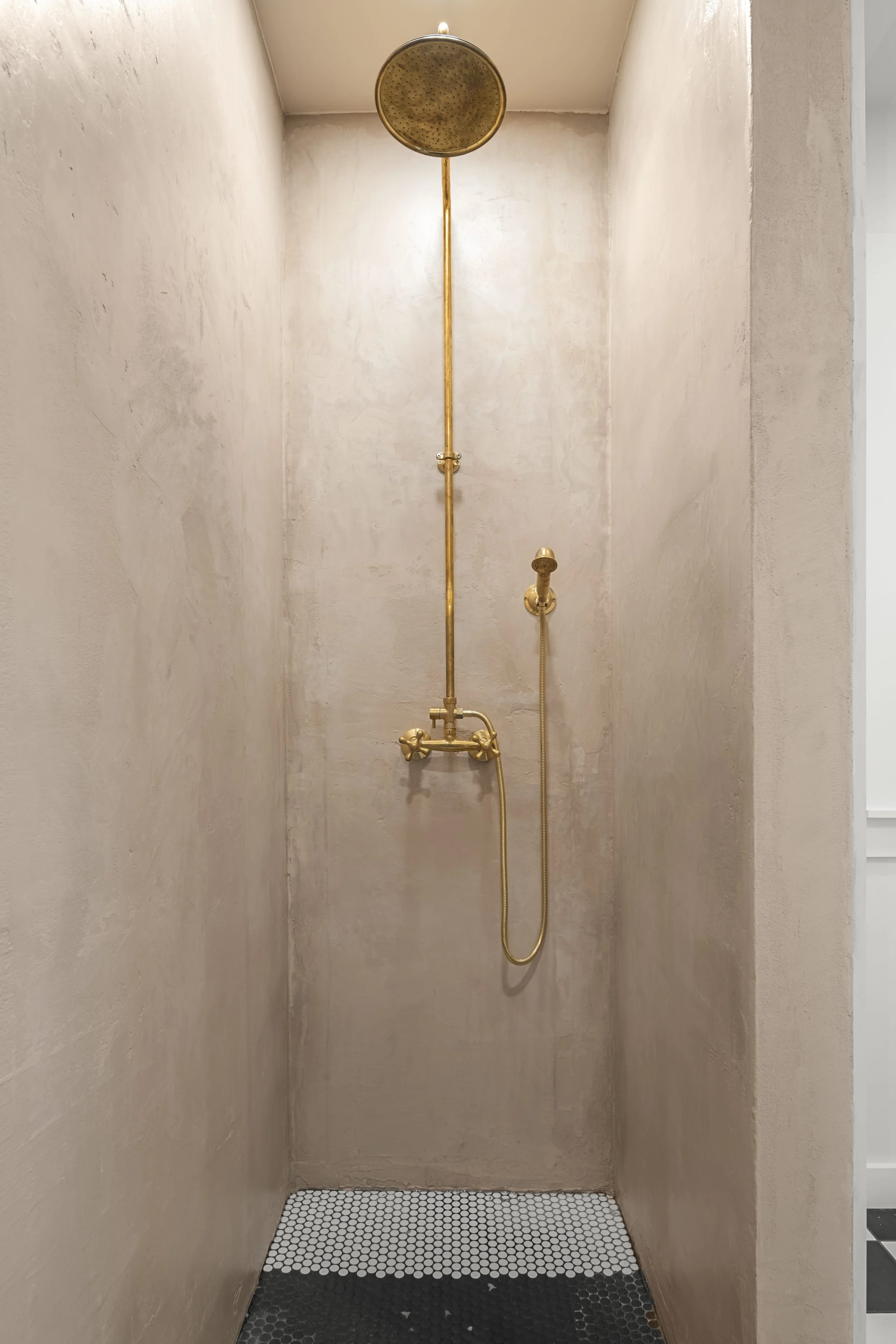 Gold-colored rainfall showerhead and handheld shower in a minimalist shower with beige walls and black-and-white hexagon tile on the floor.