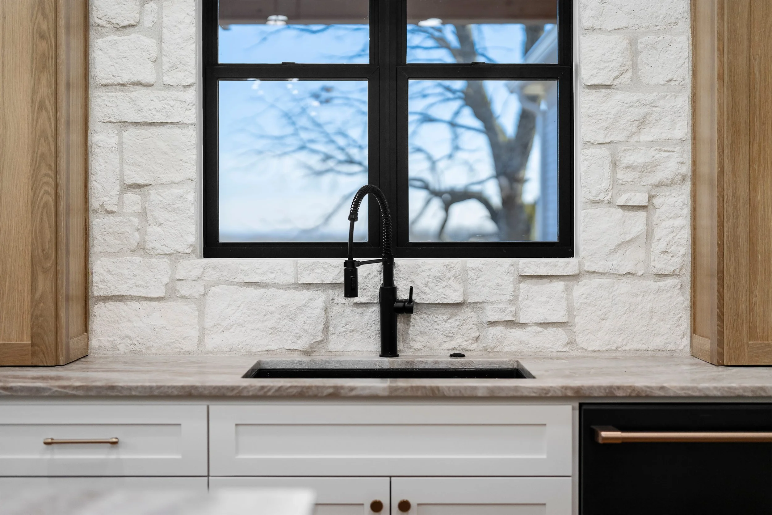 A kitchen countertop with a black faucet and sink, white cabinets with gold handles, a white brick wall, and a window showing a tree outside with snow.