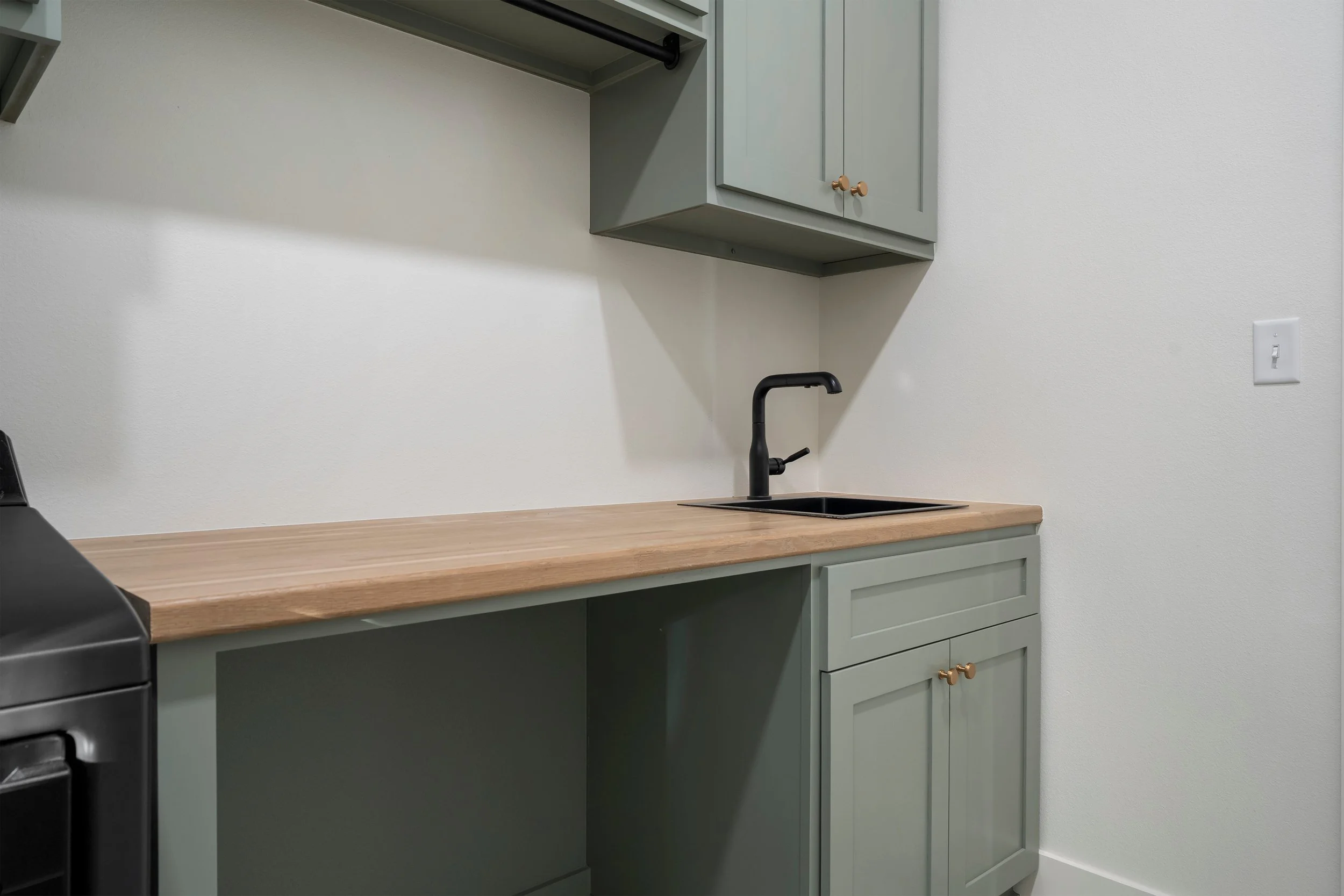 A small kitchenette with pale green cabinets, a black faucet, black sink, wooden countertop, and a wall outlet on the right.